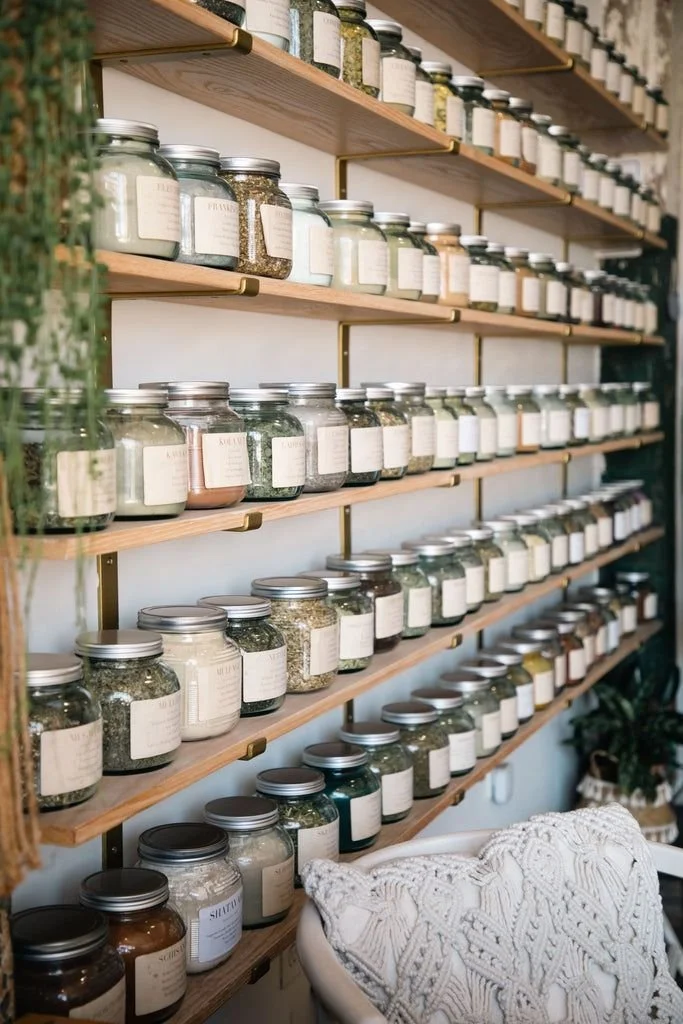 Shelves filled with glass jars containing herbs, spices, or dried goods, labeled and organized in a store or pantry setting.