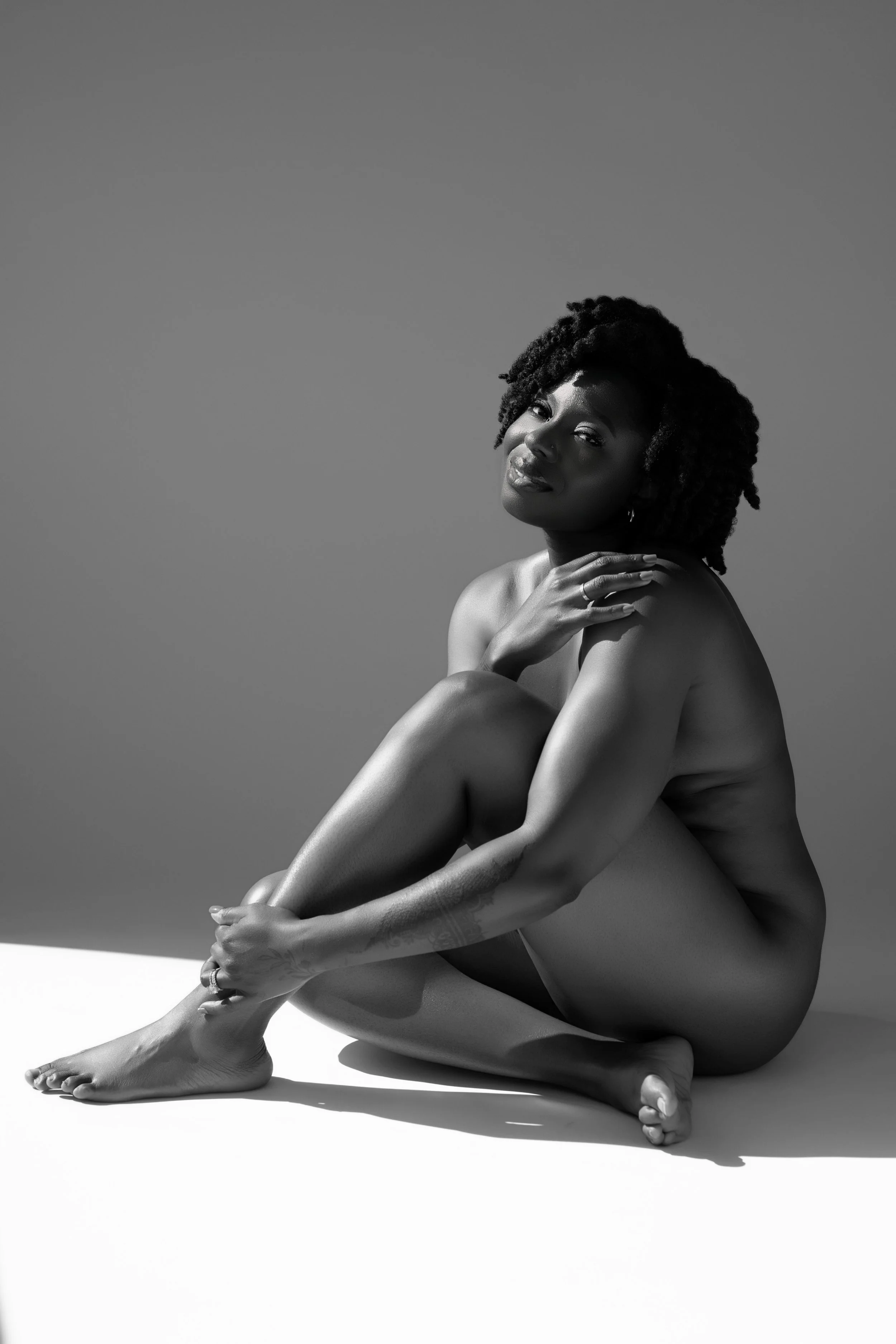 Black and white photo of a nude woman sitting with one arm around her legs, looking at the camera with a serene expression.