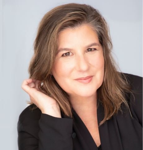 A woman with shoulder-length brown hair, smiling and wearing a black top, resting her head on her hand.