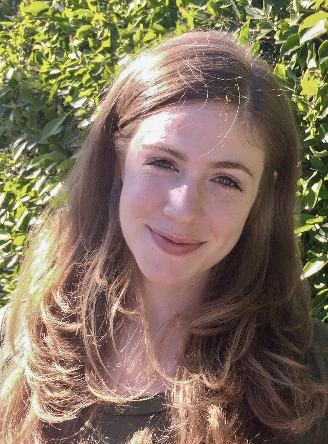 A young woman with long wavy hair taking a selfie outdoors in front of green bushes, smiling at the camera in bright sunlight.