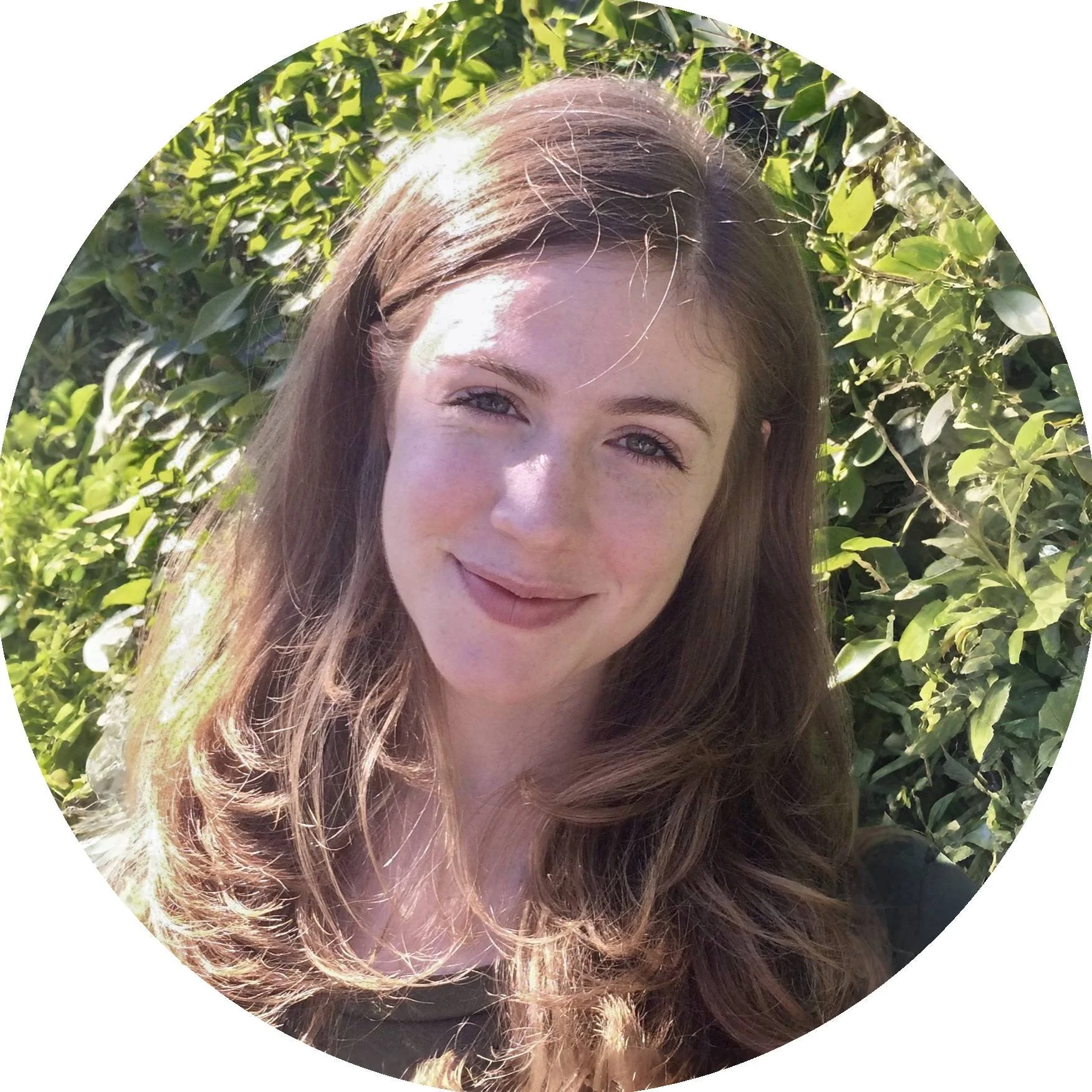 Alice O'Connor, a woman with long, wavy brown hair smiling outdoors next to green foliage, sunlight on her face.