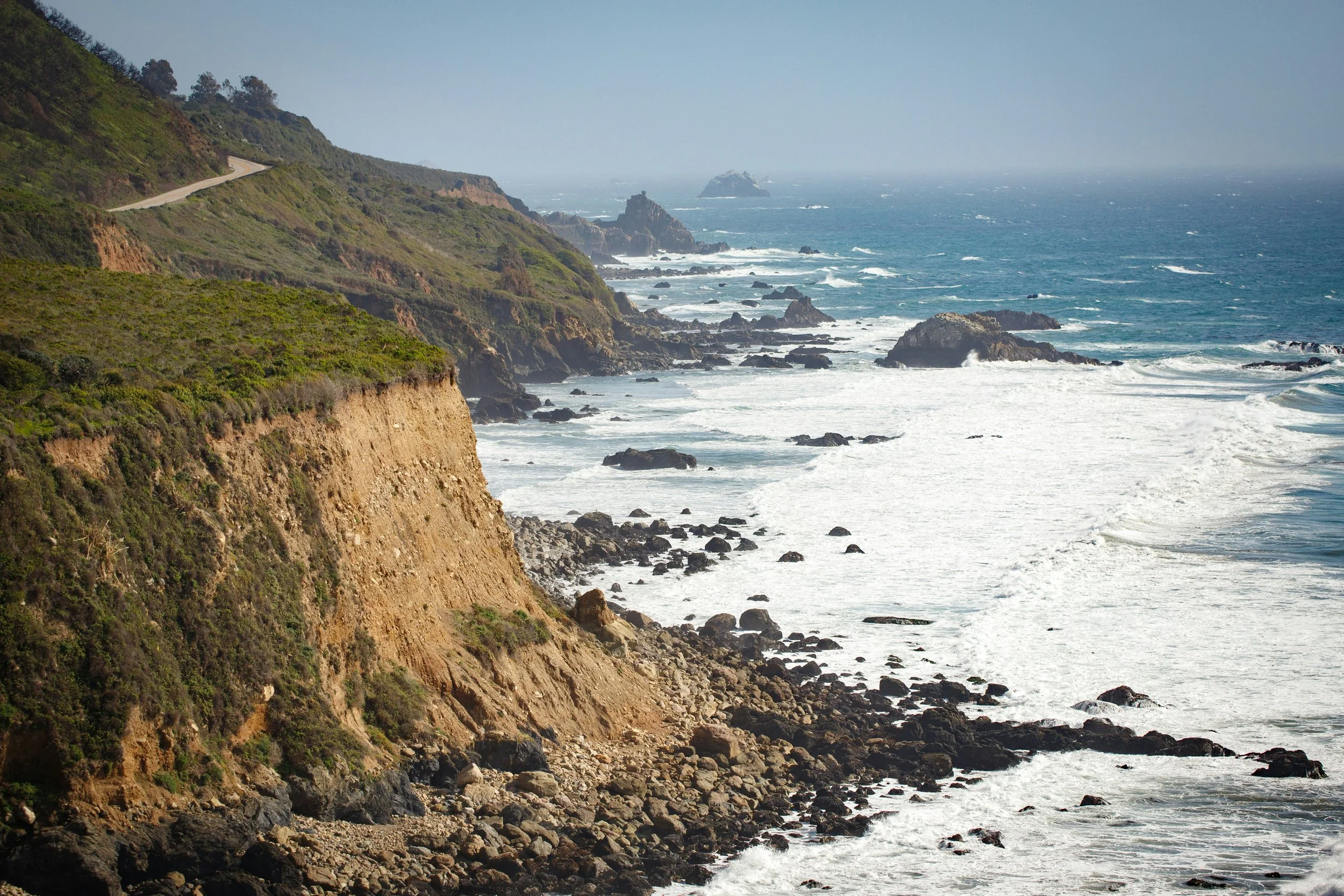 Coastal cliffs with greenery on top, rocky shoreline below, and ocean waves crashing against rocks in the distance under a clear sky.