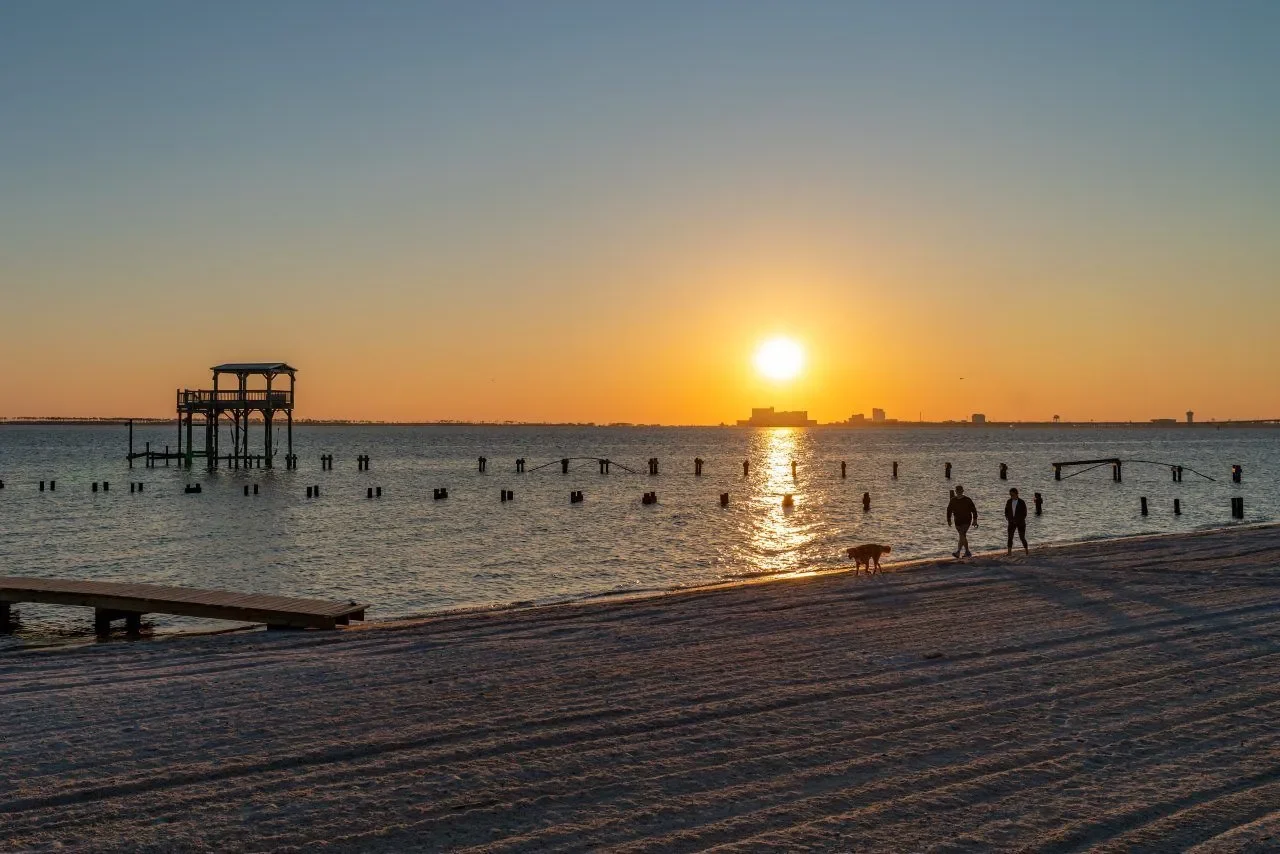 Sunset over a beach with two people walking a dog along the shoreline, a pier in the water, and buildings in the distance.