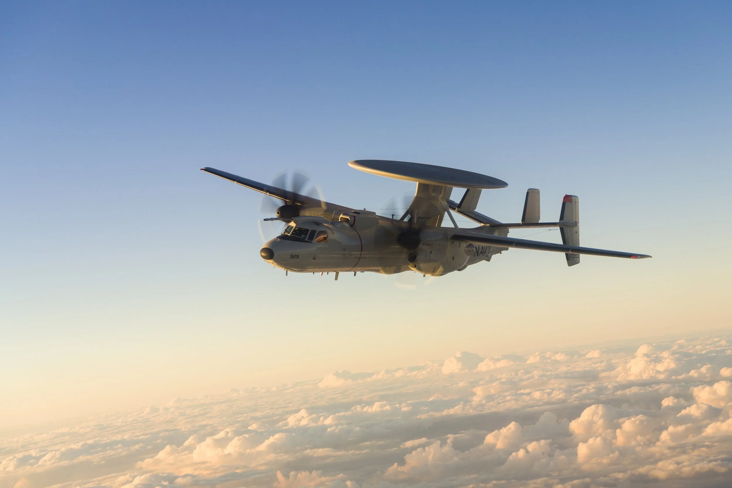 A Navy aircraft flying above clouds during sunset or sunrise.