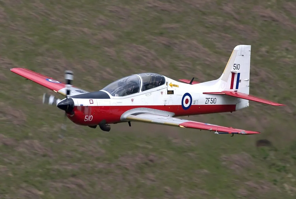 A small propeller aircraft flying over a grassy hillside, painted in red, white, and blue with military markings.