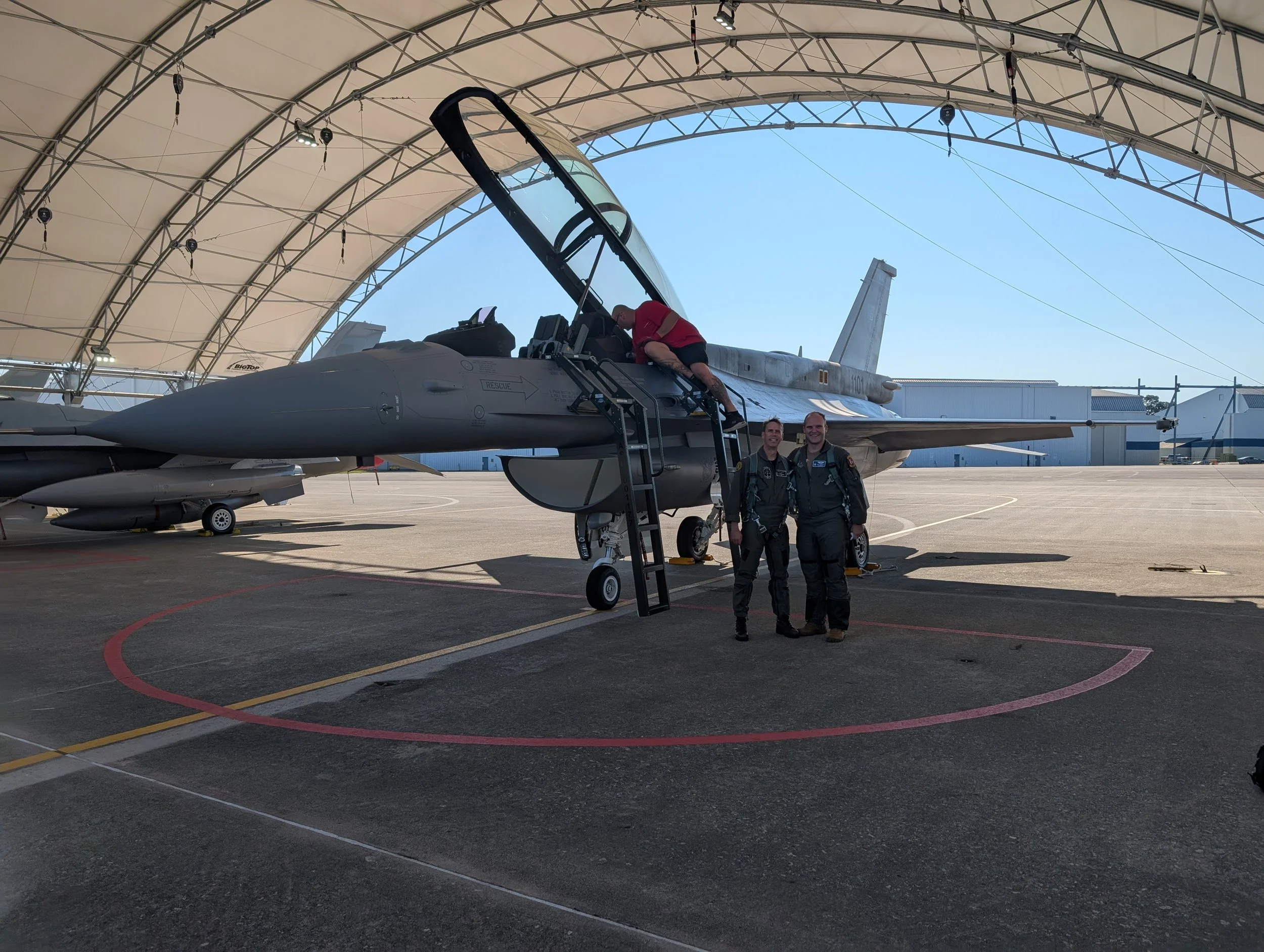 Two uniformed pilots standing on tarmac beside a F-16 fighter jet inside a large hangar, with one person climbing into the cockpit.