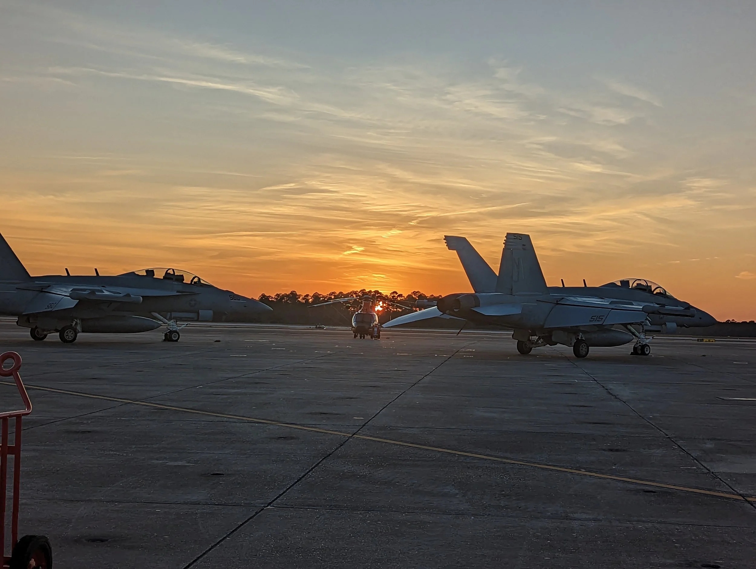 Two fighter jets on an airport tarmac at sunset with a small vehicle between them, and a line of trees in the background.
