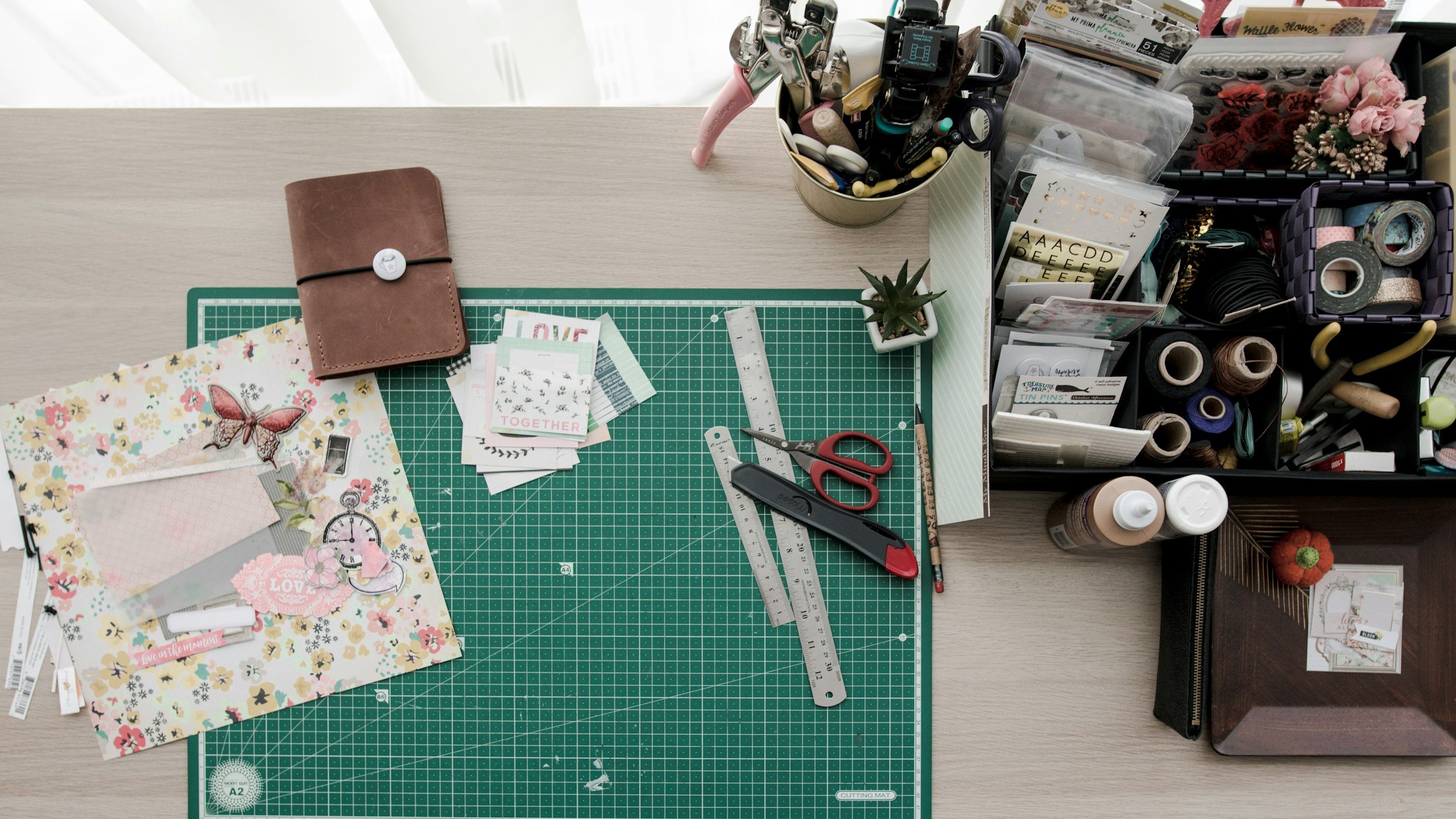A crafting workspace with a green cutting mat, scissors, rulers, and various decorative paper and stickers, along with storage containers filled with washi tapes, flowers, and craft supplies.
