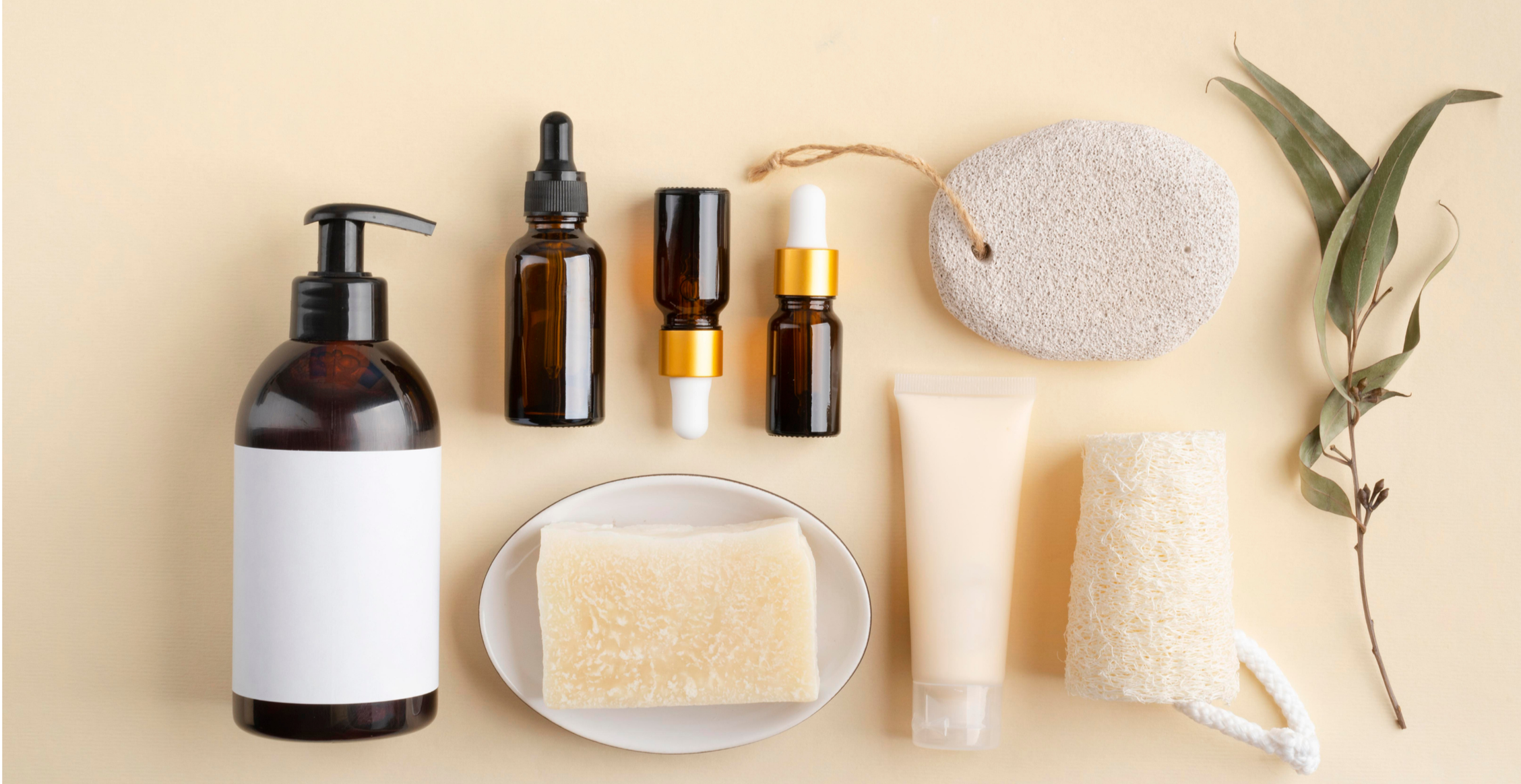 Skincare and beauty products arranged on a beige background, including a pump bottle, small dropper bottles, a soap bar on a dish, a tube, a textured sponge, and natural elements like a stone and a branch with leaves.