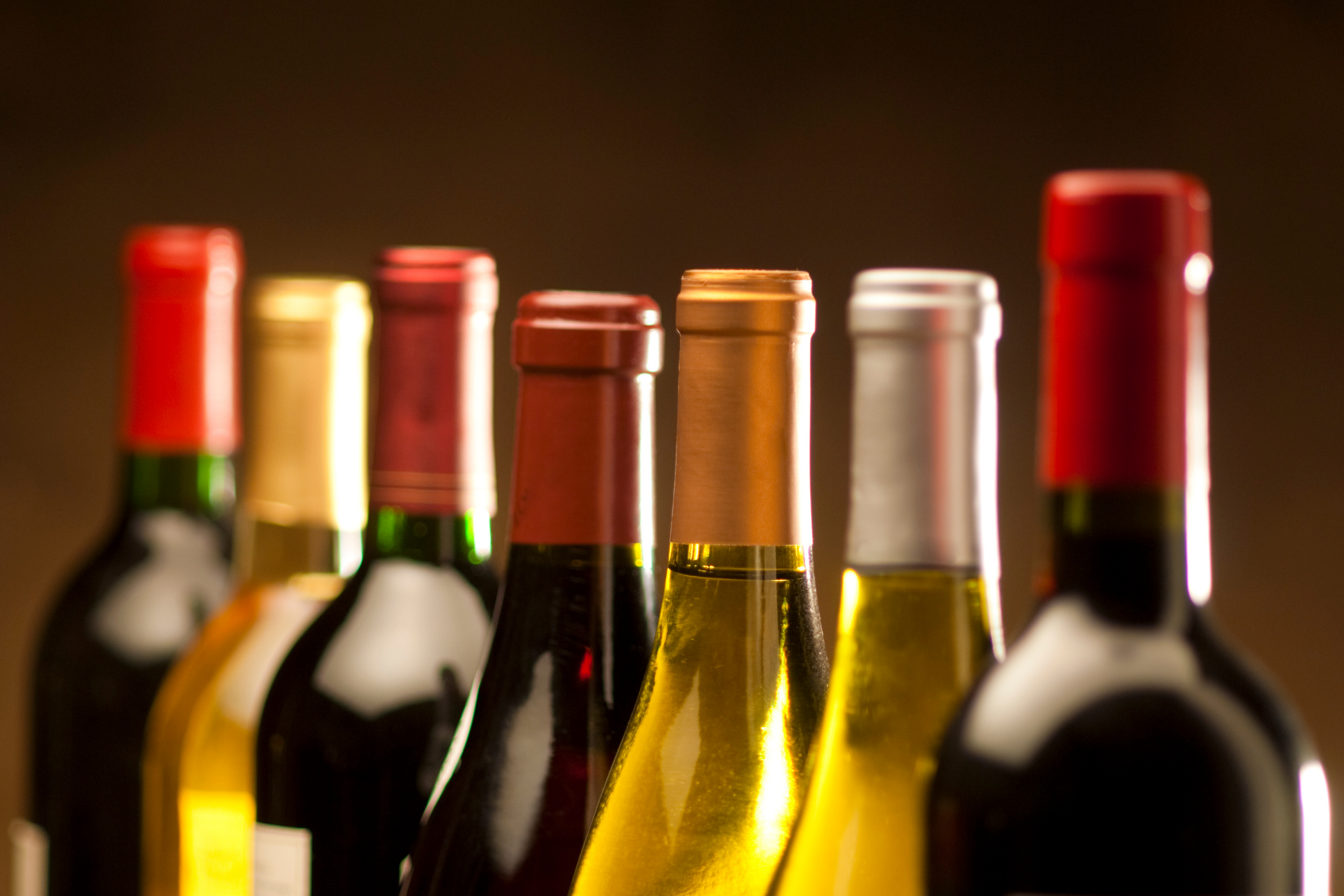 Close-up of various bottles of wine and champagne with colorful caps, illuminated against a dark background.