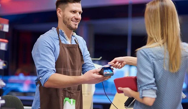 A man wearing a brown apron is scanning a credit card or smartphone for payment from a woman at a register in a store or cafe.