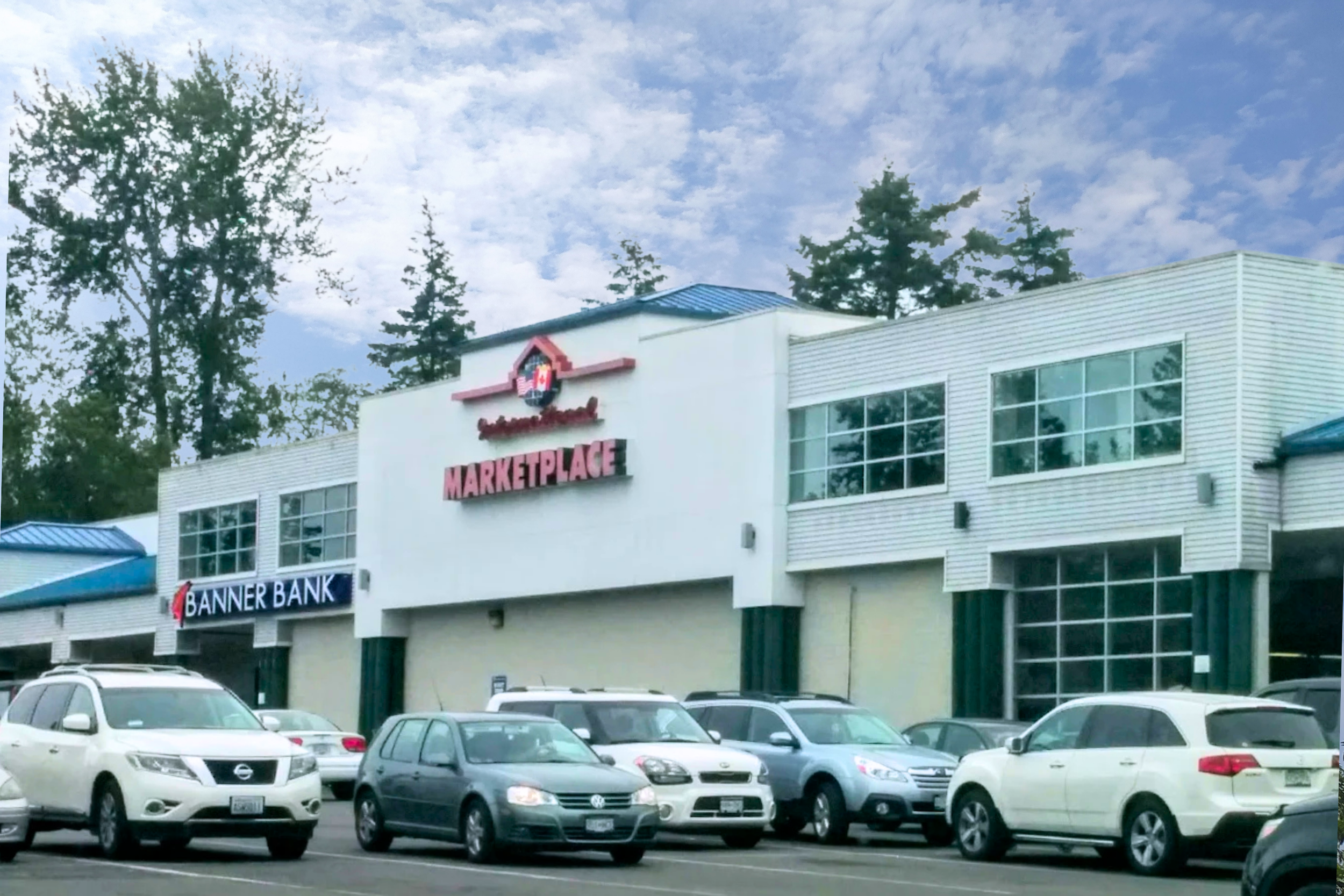 Shopping mall with storefront signs for Banner Bank and Marketplace, parked cars in parking lot, and trees with a partly cloudy sky
