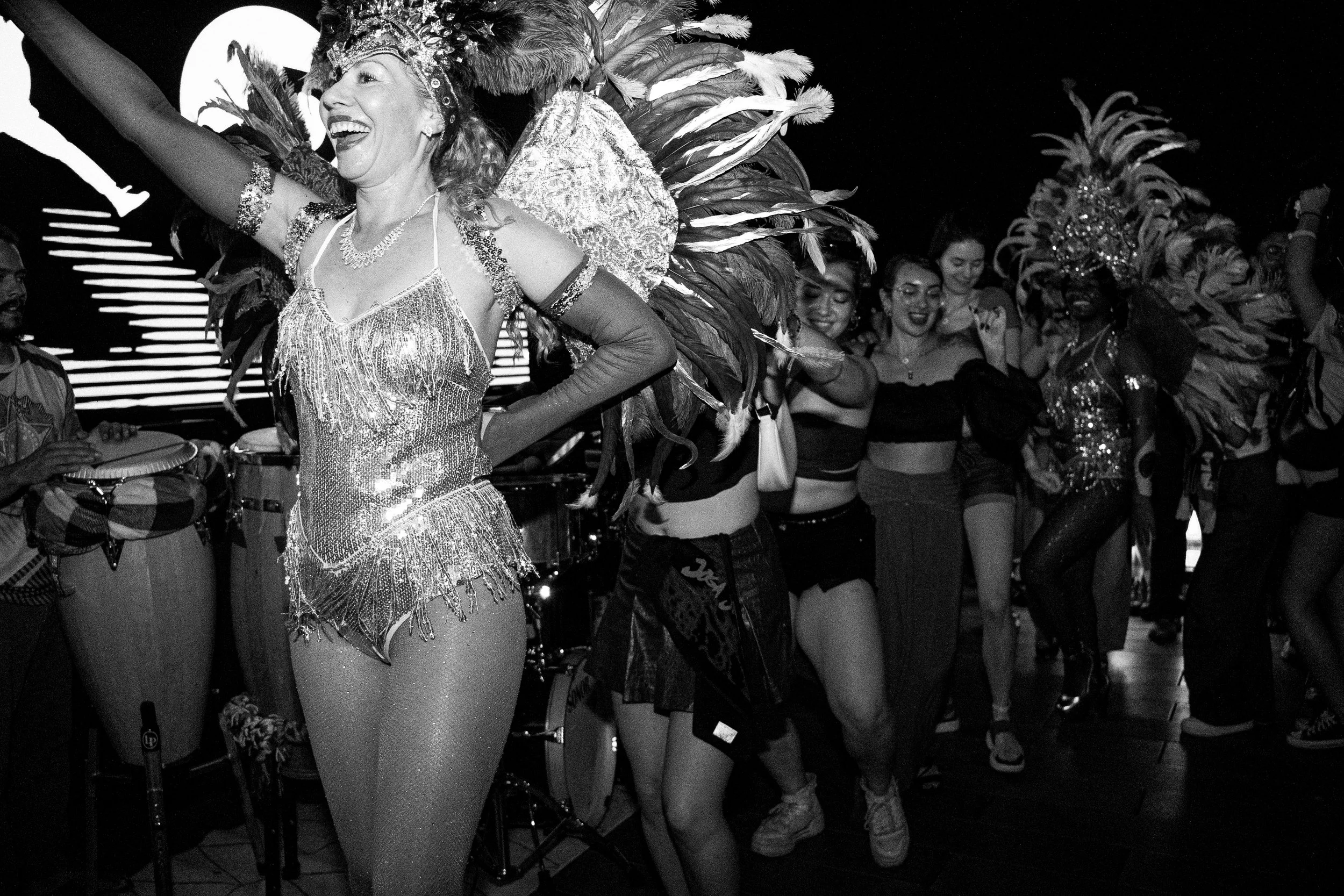 A woman dressed in a sparkling, fringed costume with feathers on her head, smiling and dancing in a parade. She is surrounded by other women in similar costumes and a crowd of people, some taking photos.