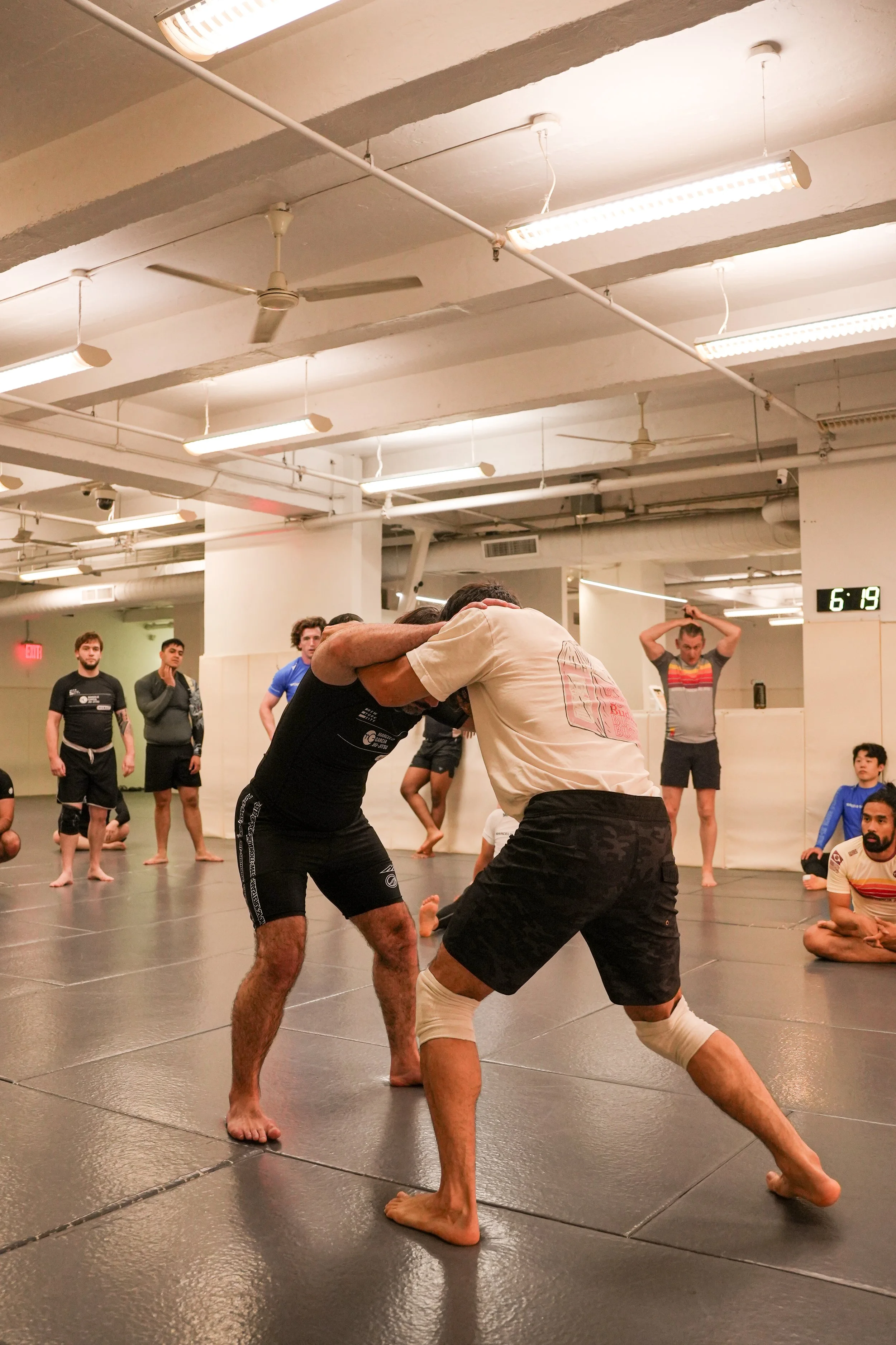 Two men practicing Brazilian Jiu-Jitsu in a martial arts gym, wearing rash guards, gripping each other and engaged in a grappling technique, while others watch in the background.