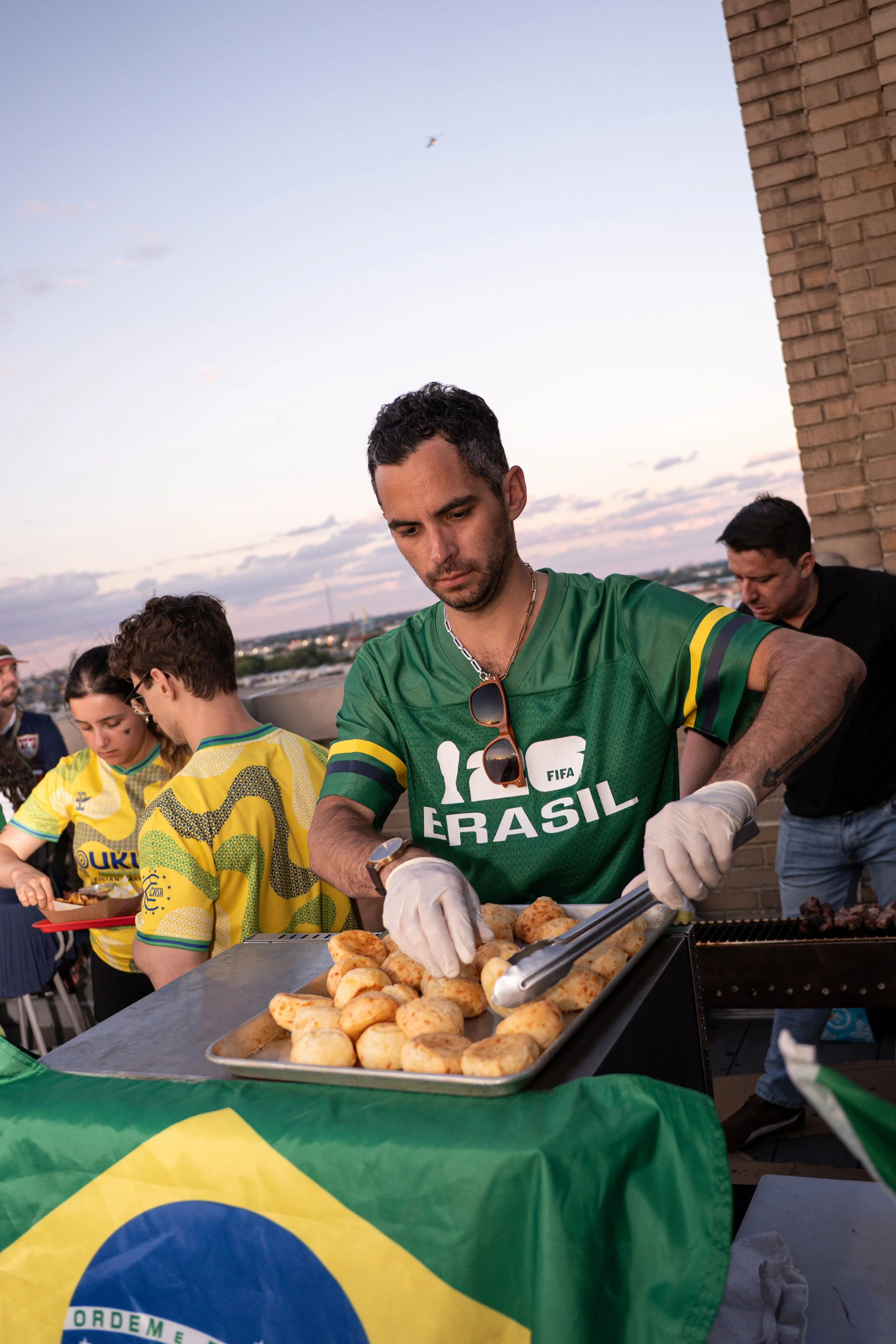 A man in a green Brazil national football team shirt is serving food at an outdoor event, with several other people around him. The man is wearing gloves and sunglasses hanging from his shirt. A large Brazilian flag is draped over the table. The back