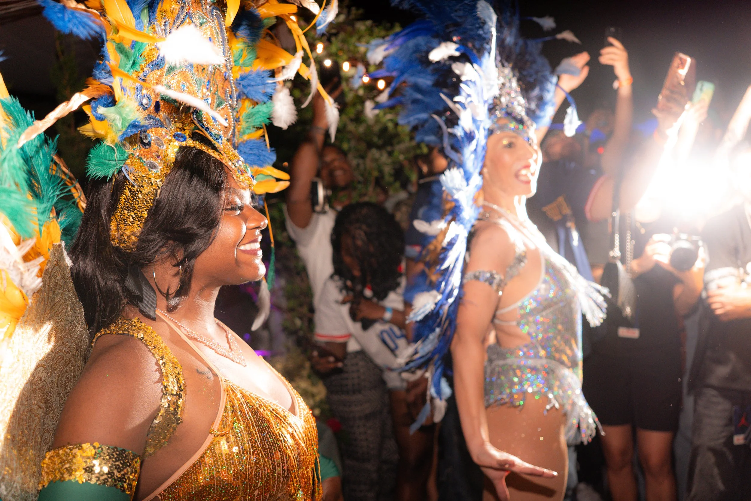 People wearing colorful, elaborate costumes with feathered headpieces, celebrating at a festive event at night.