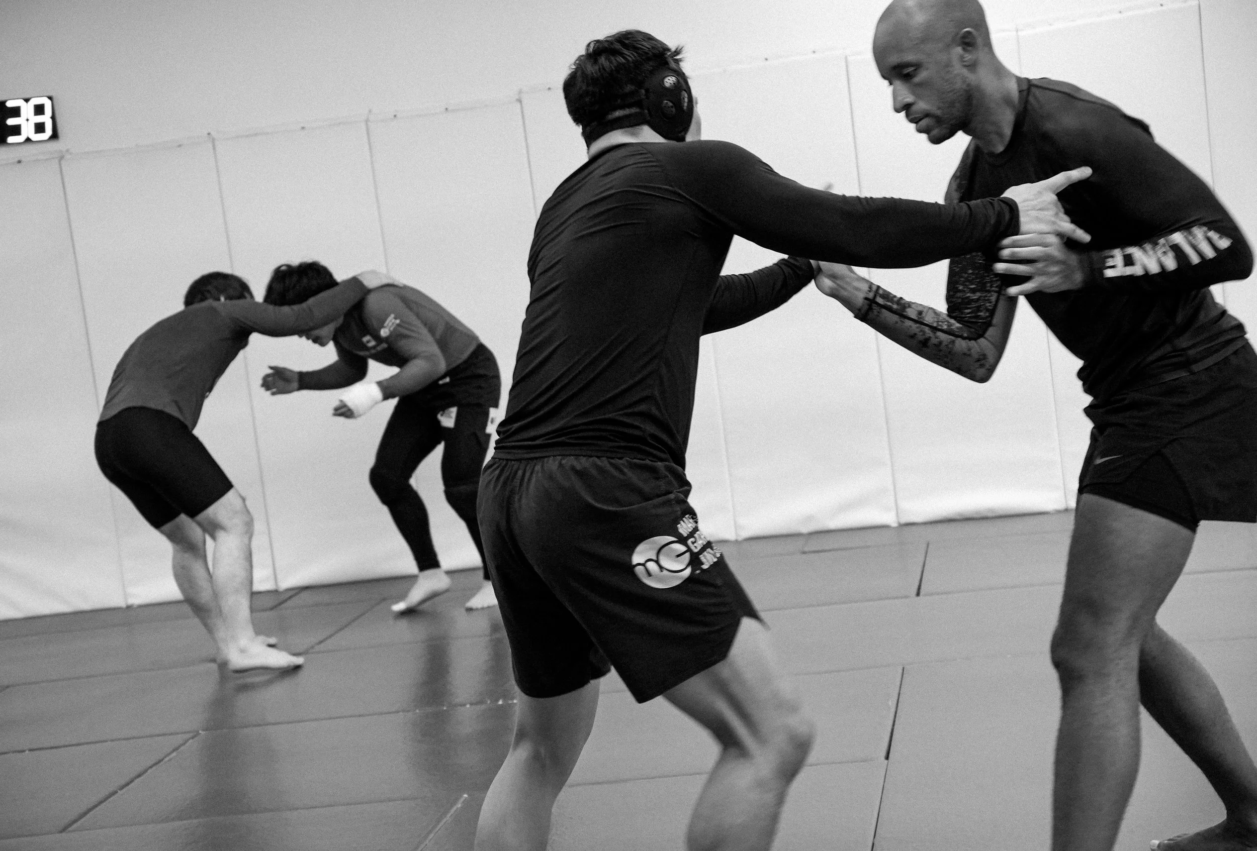 Black and white photo of martial arts students practicing with partners in a gym, wearing rash guards and shorts, with a timer displaying 38 seconds on the wall.