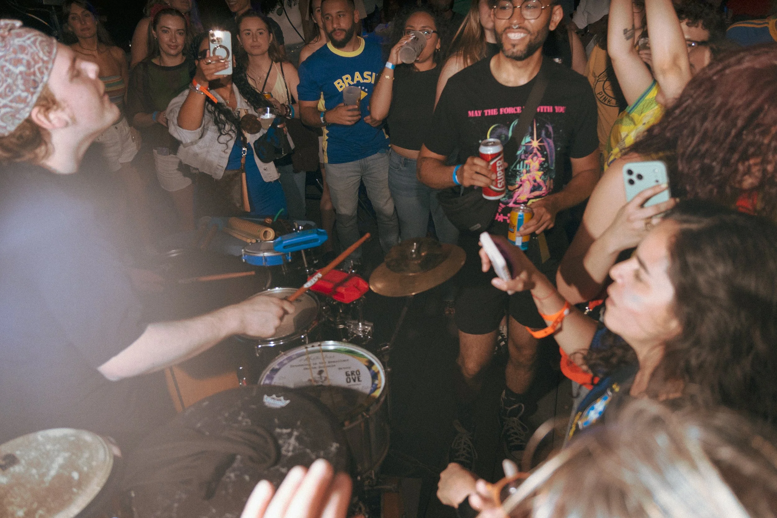 A crowd of people at a party, with some holding drinks and taking photos, gathered around a drum set, with a person playing the drums in the foreground.