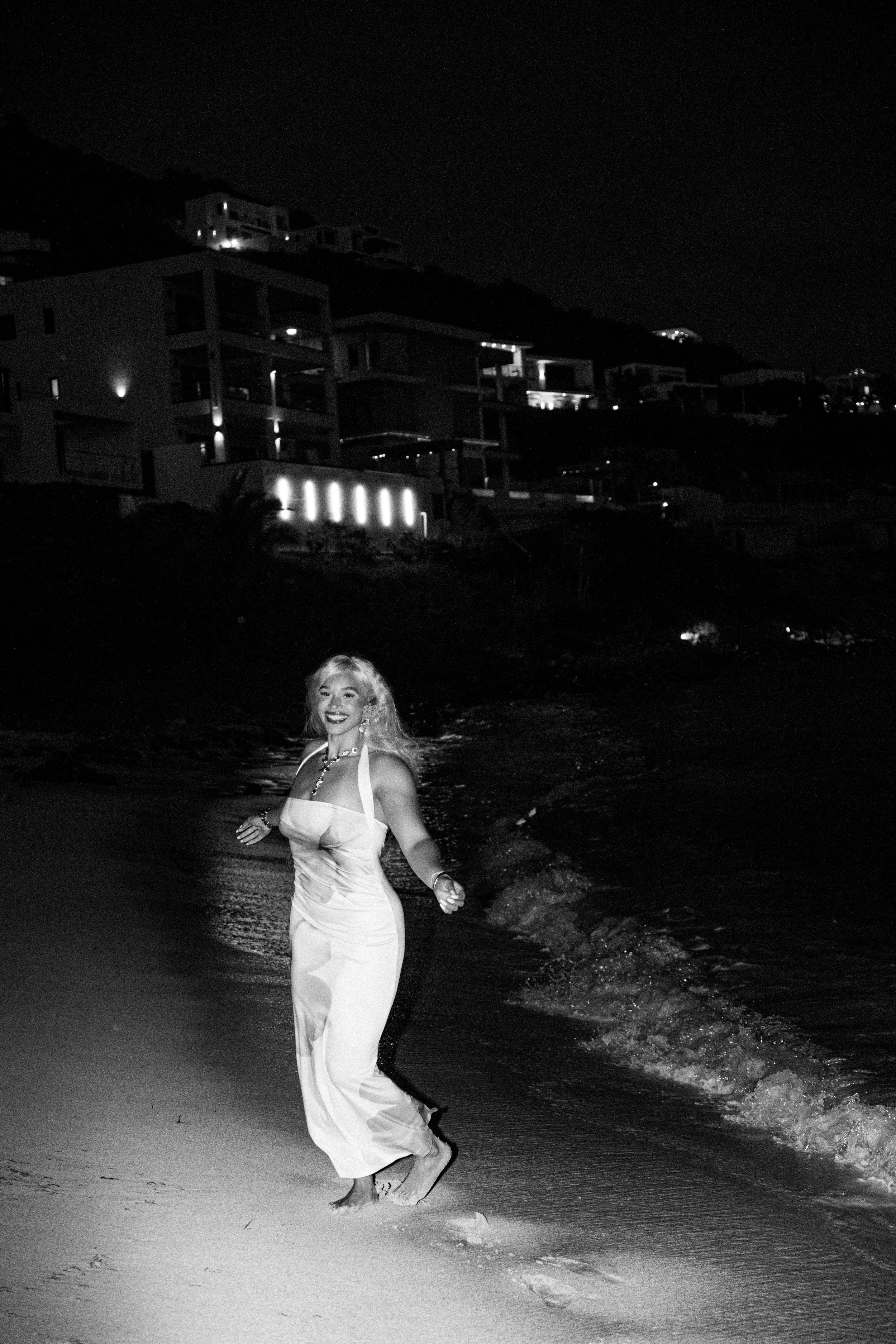 A woman in a white dress smiling and walking barefoot on the beach at night, with buildings lit up on a hillside in the background.