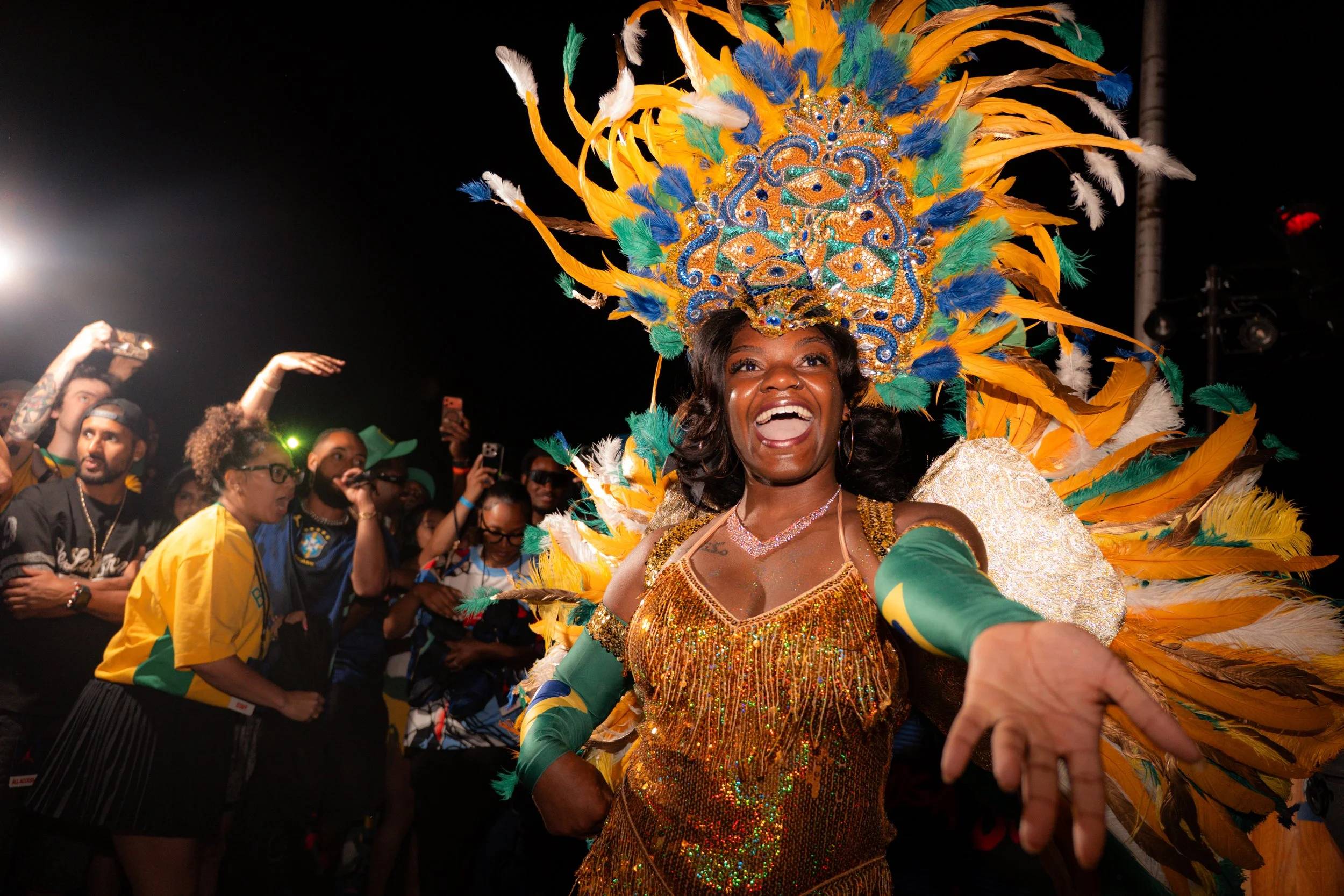 A woman dressed in a vibrant, sequined costume with a large feathered headdress, smiling and dancing at a nighttime celebration with a crowd around her.