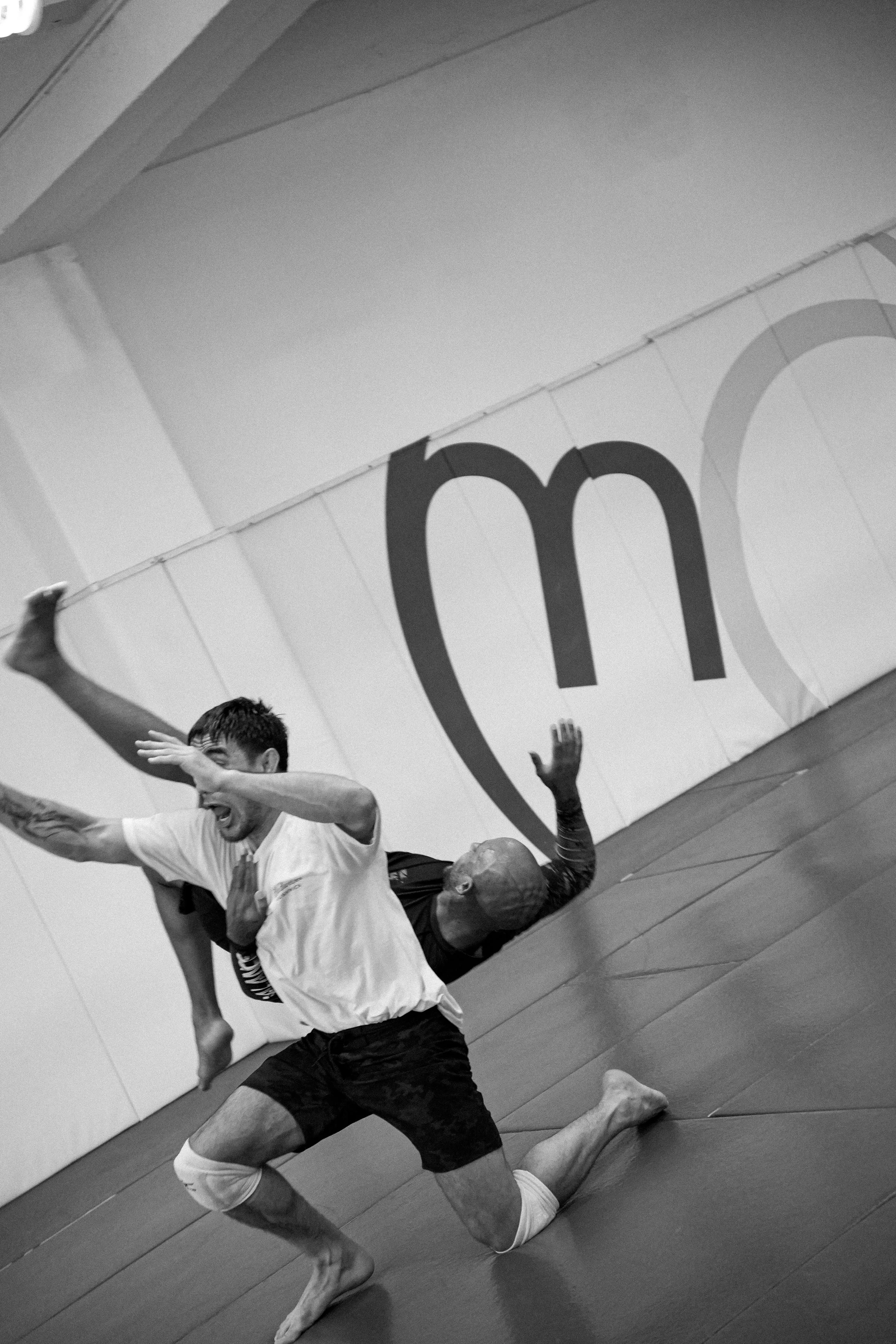 Two men practicing Brazilian Jiu-Jitsu on a padded floor. One man, wearing a white T-shirt and shorts, is executing a submission move on another man, who is bald, tattooed, and wearing dark shorts. A large banner with partial text is visible in the b