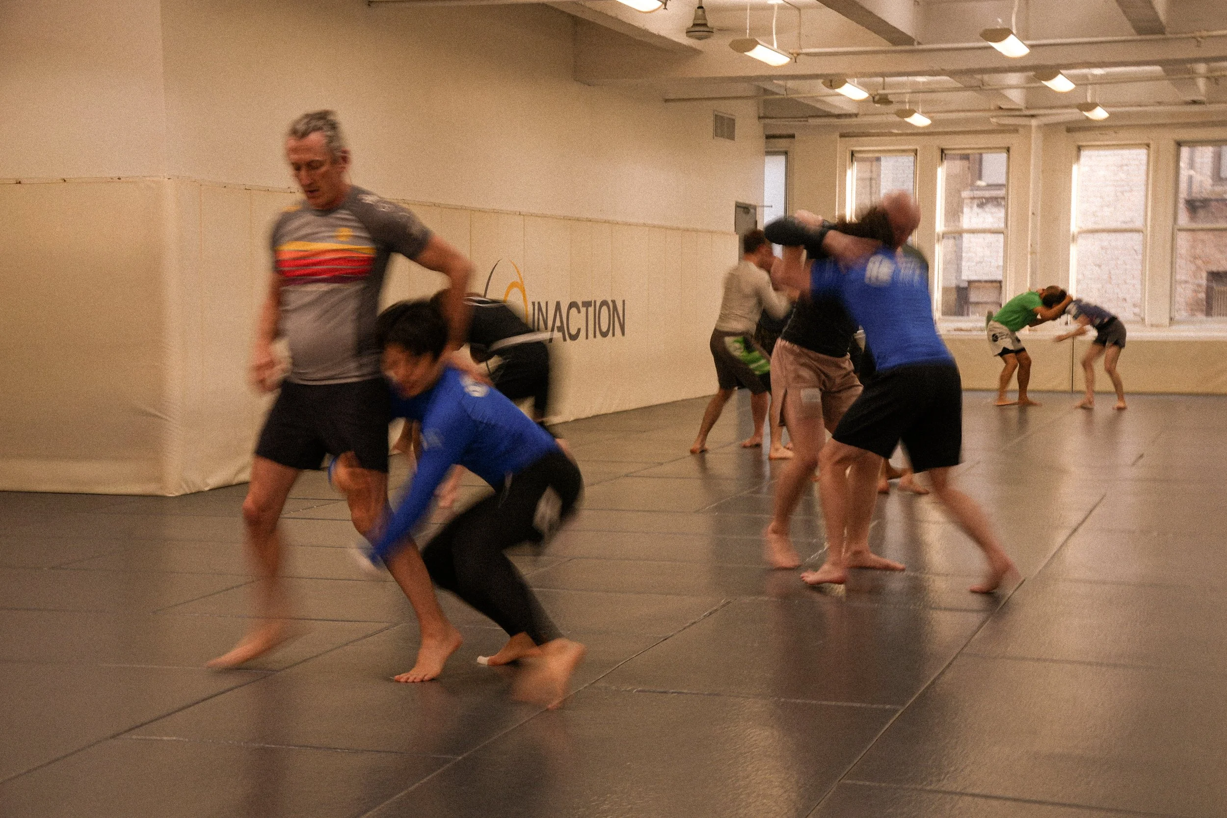 People practicing Brazilian Jiu-Jitsu in a training room with mats, some grappling and some observing, with windows in the background.
