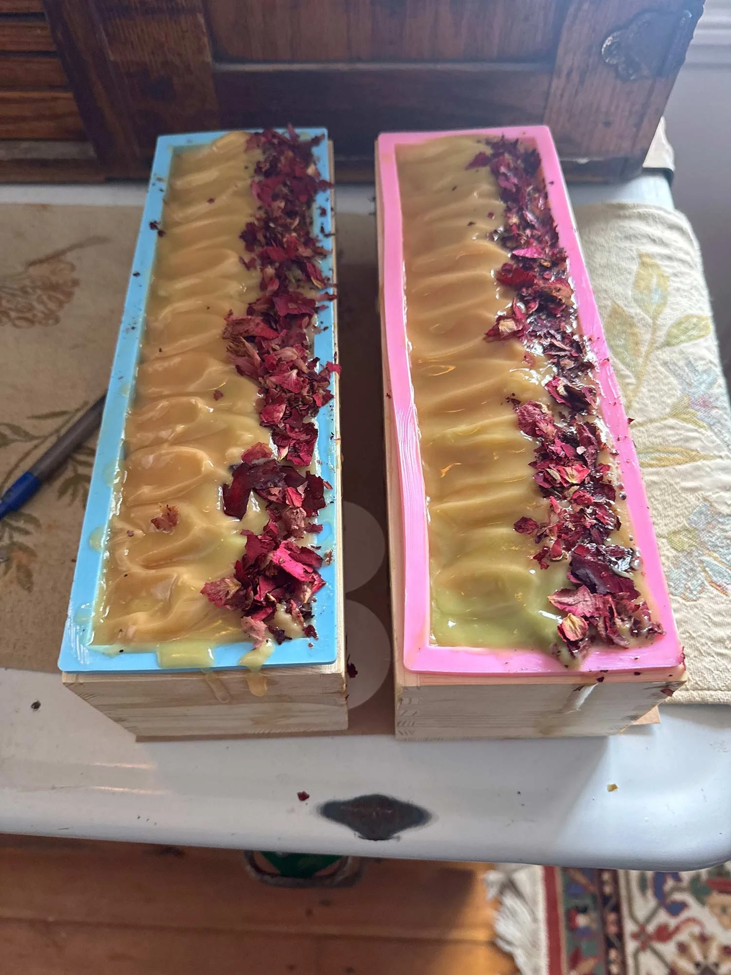 Two soap molds filled with pale yellow soap and topped with dried rose petals, one with a blue border and the other with a pink border.