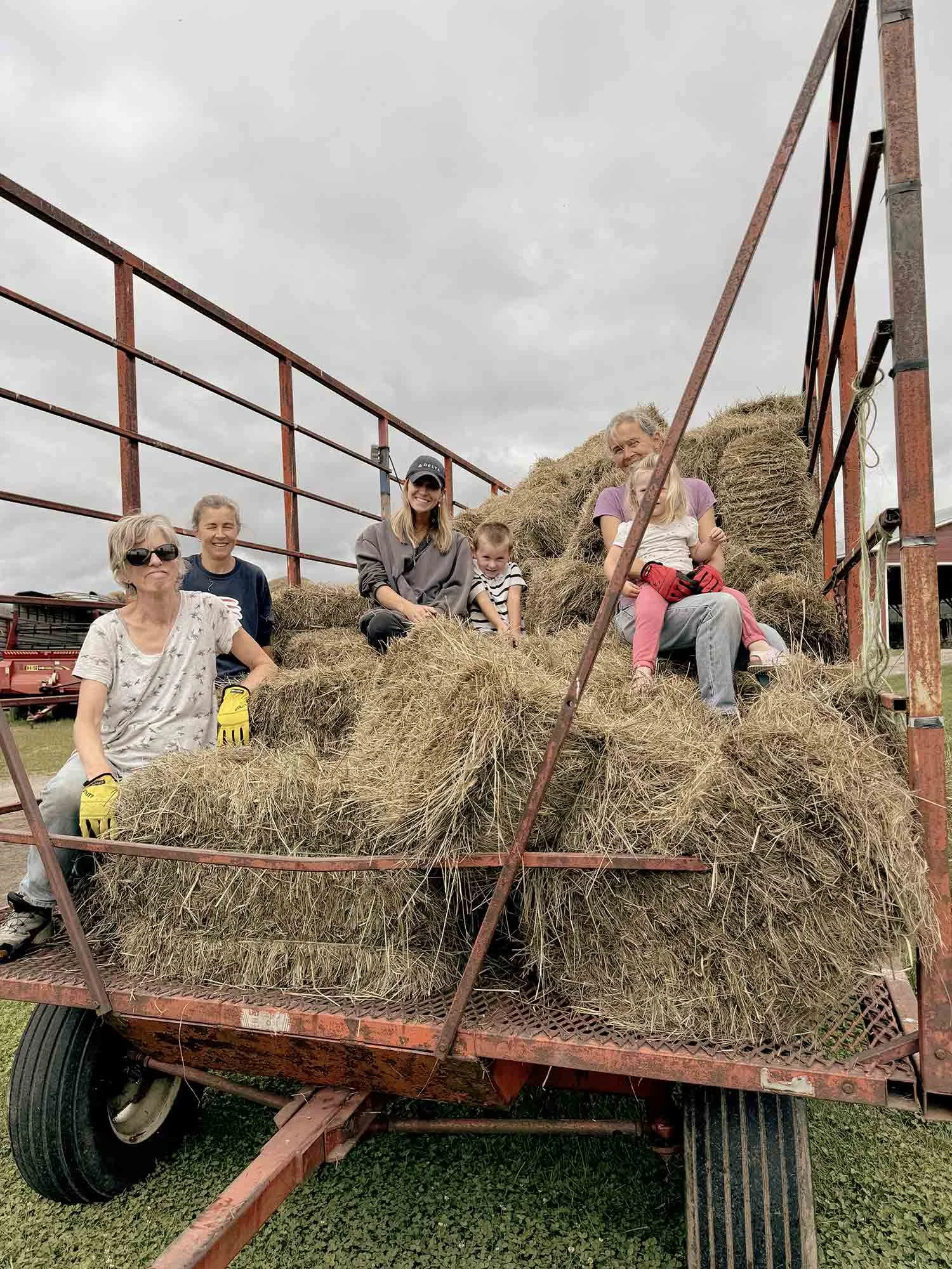 Group of five people, including children, sitting on a hay wagon filled with hay bales on a farm.