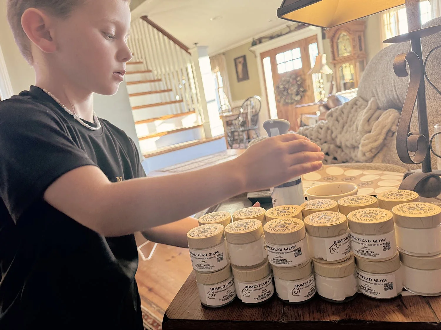 A young boy in a black shirt arranging jars of homemade glow body butter on a wooden table in a cozy, well-lit kitchen or dining room.