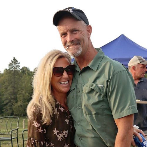 A smiling man and woman, standing outdoors, with trees and a blue tent in the background, at a casual outdoor gathering.