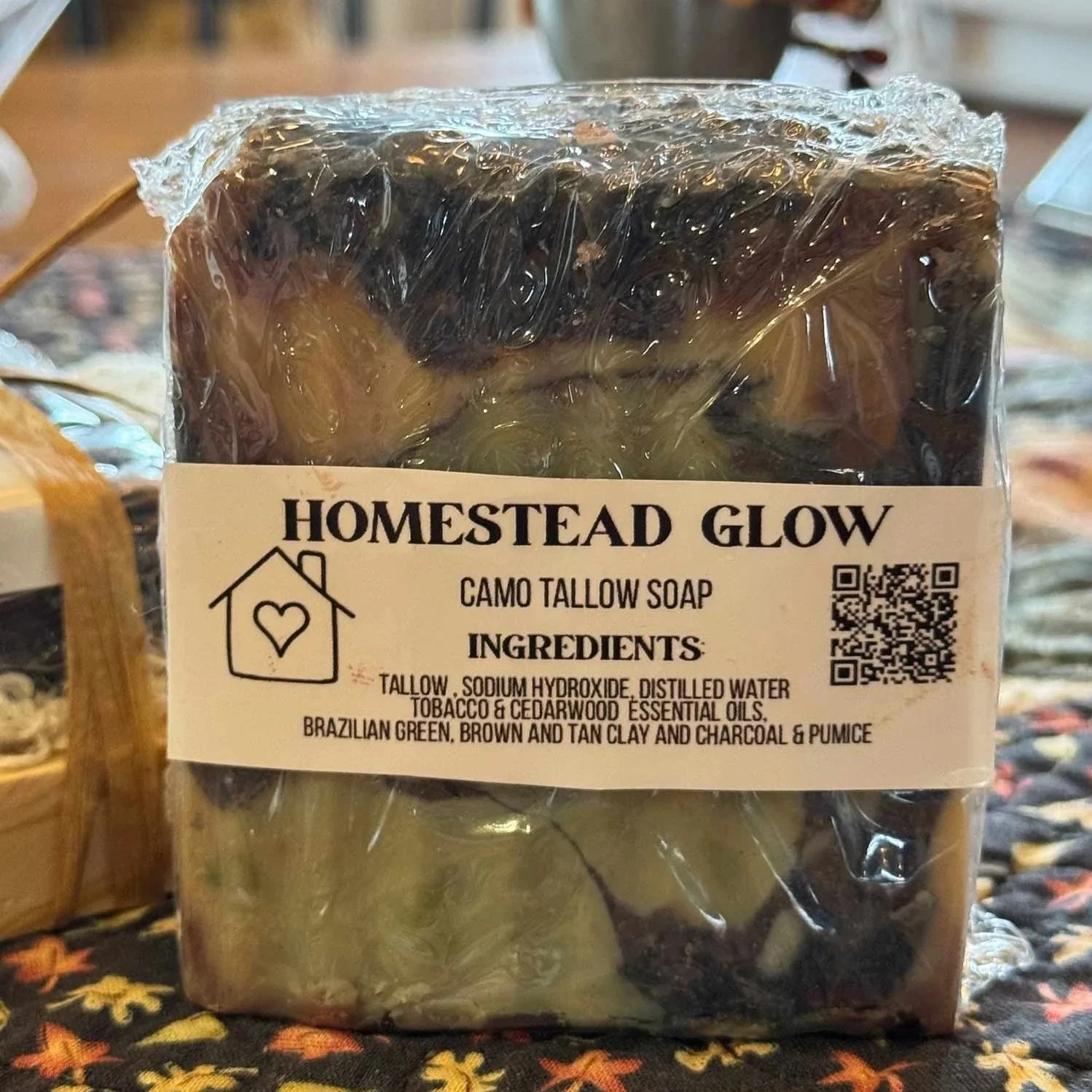 A bar of soap labeled 'Homestead Glow Camo Tallow Soap' with natural ingredients listed on the front, placed on a colorful patterned fabric surface.
