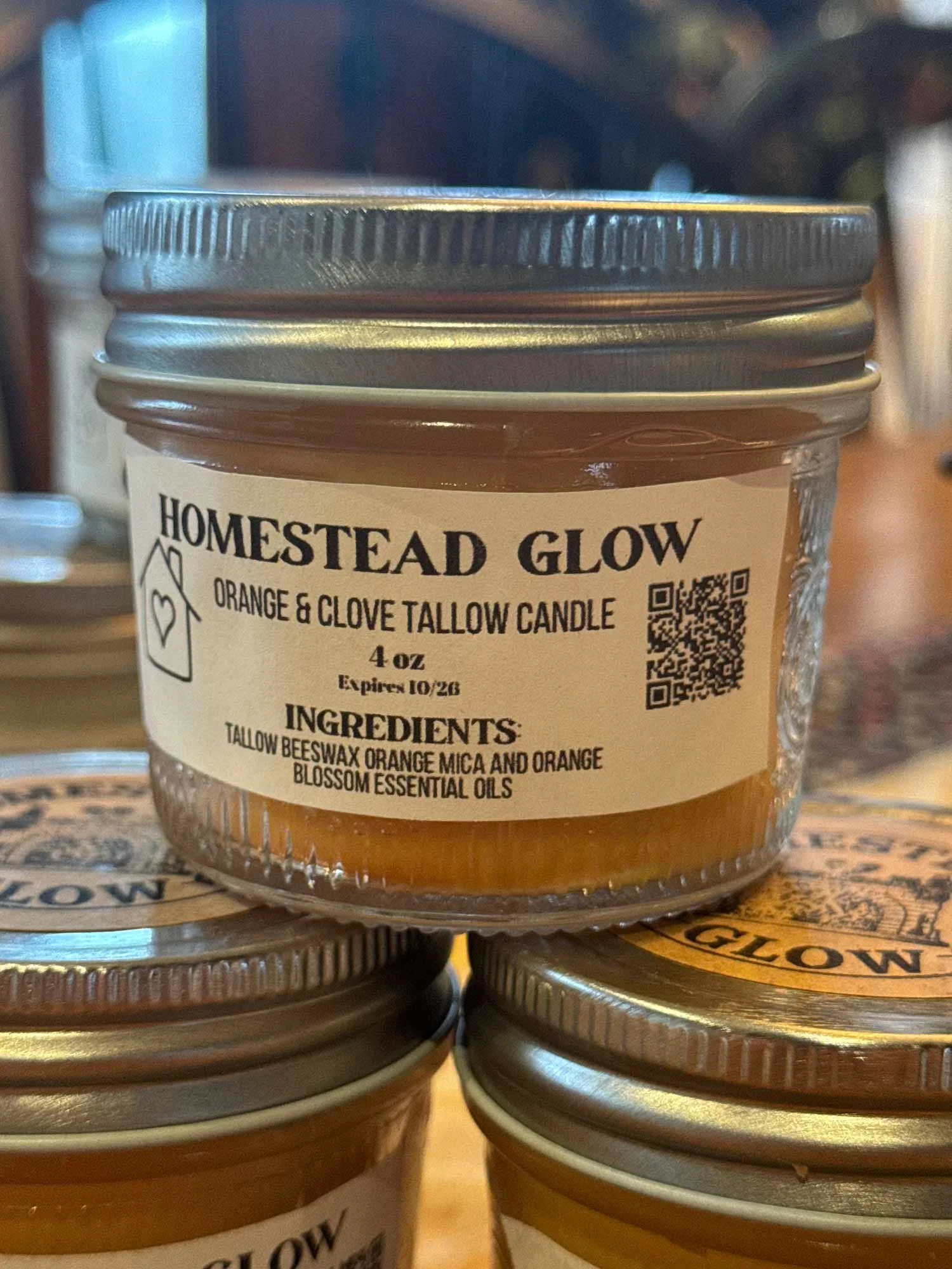 Close-up of a jar labeled 'Homestead Glow' orange and clove tallow candle, with ingredients listed as tallow, beeswax, orange mica, and orange blossom essential oils.