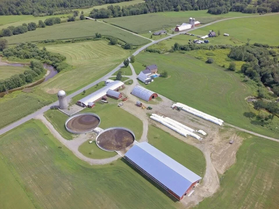 Aerial view of a farm with multiple barns, silos, green fields, a small pond, and farm equipment.