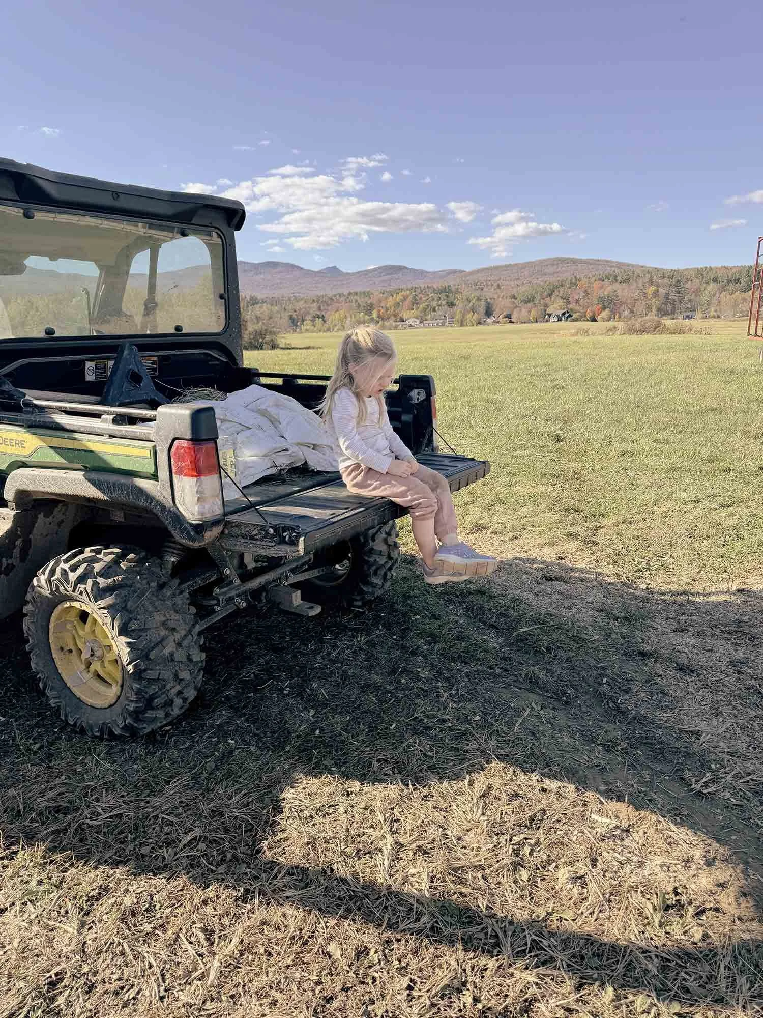 A young girl sitting on the tailgate of a green John Deere utility vehicle in a grassy field with mountains and trees in the background under a partly cloudy sky.