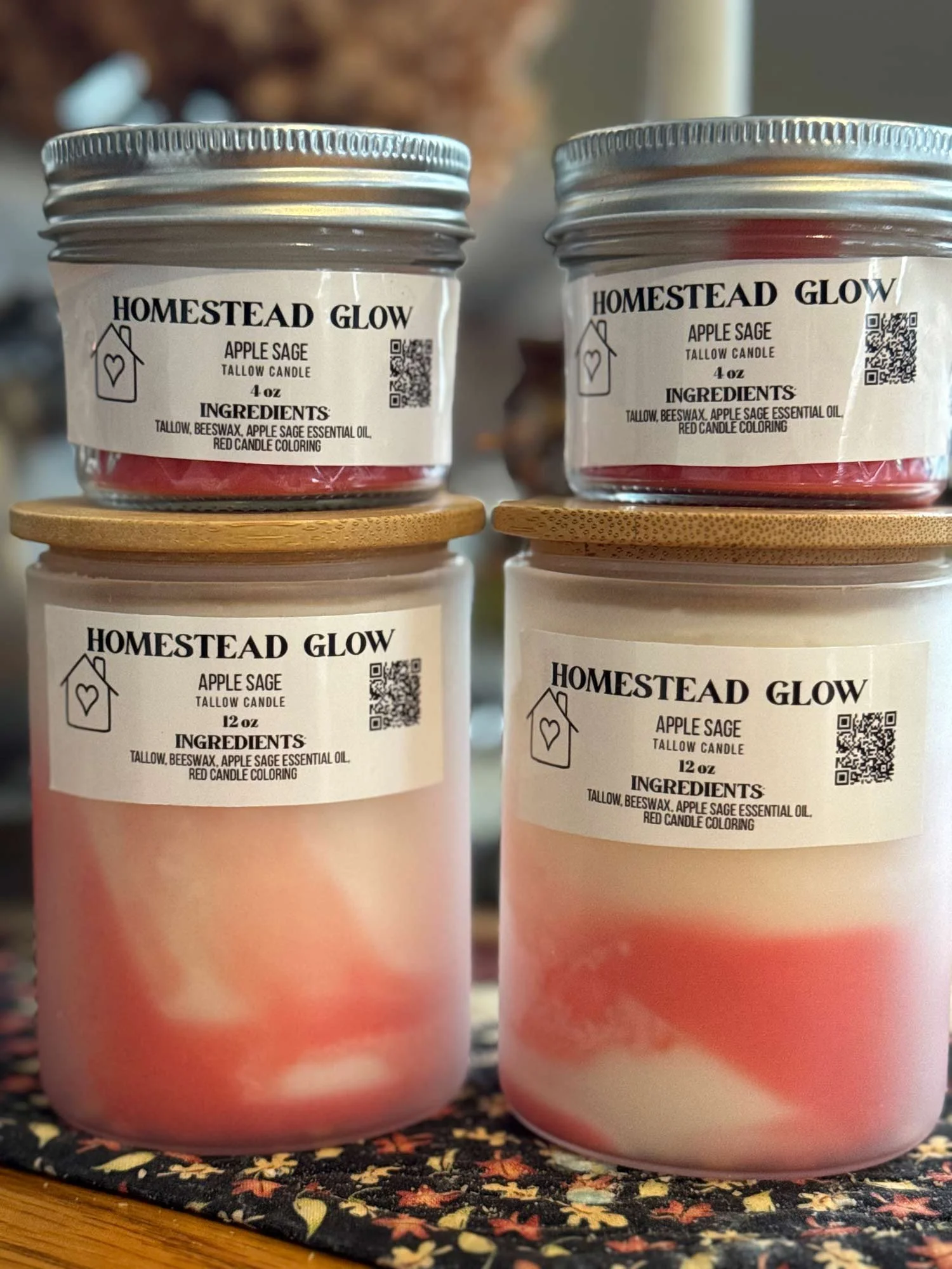 Four candles labeled 'Homestead Glow Apple Sage Tallow Candle' in different sizes, with metal and wooden lids, placed on a decorative cloth.