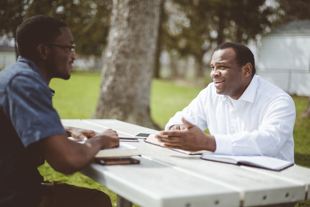 Two men sit at a picnic table outdoors, talking and smiling, with open books and notebooks in front of them.