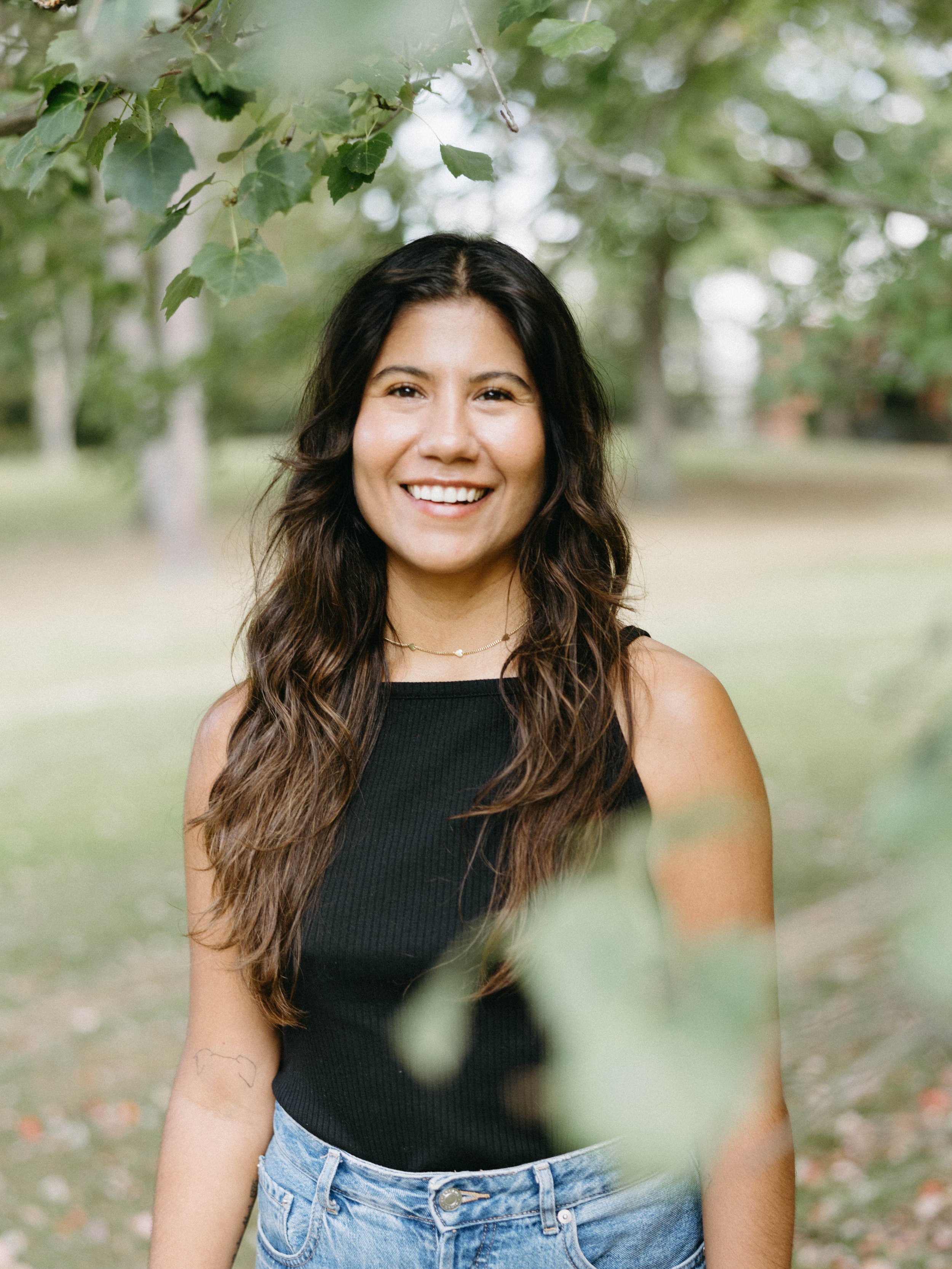 Smiling woman with wavy hair wearing a black sleeveless top and blue jeans standing outdoors under a tree with leaves.