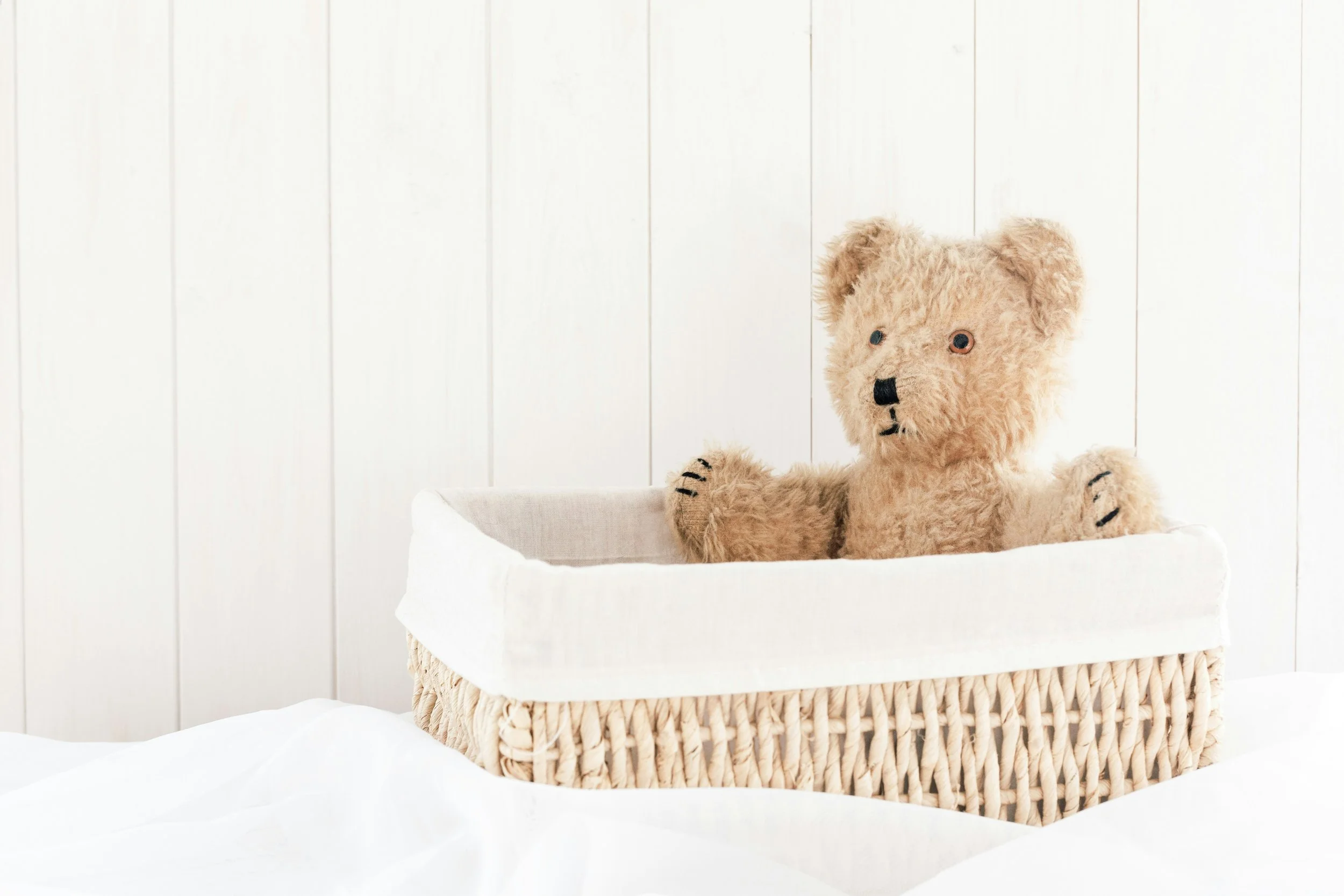 Vintage teddy bear with plastic eyes and stitched nose sitting in a woven basket on a white bed with white wall paneling background.