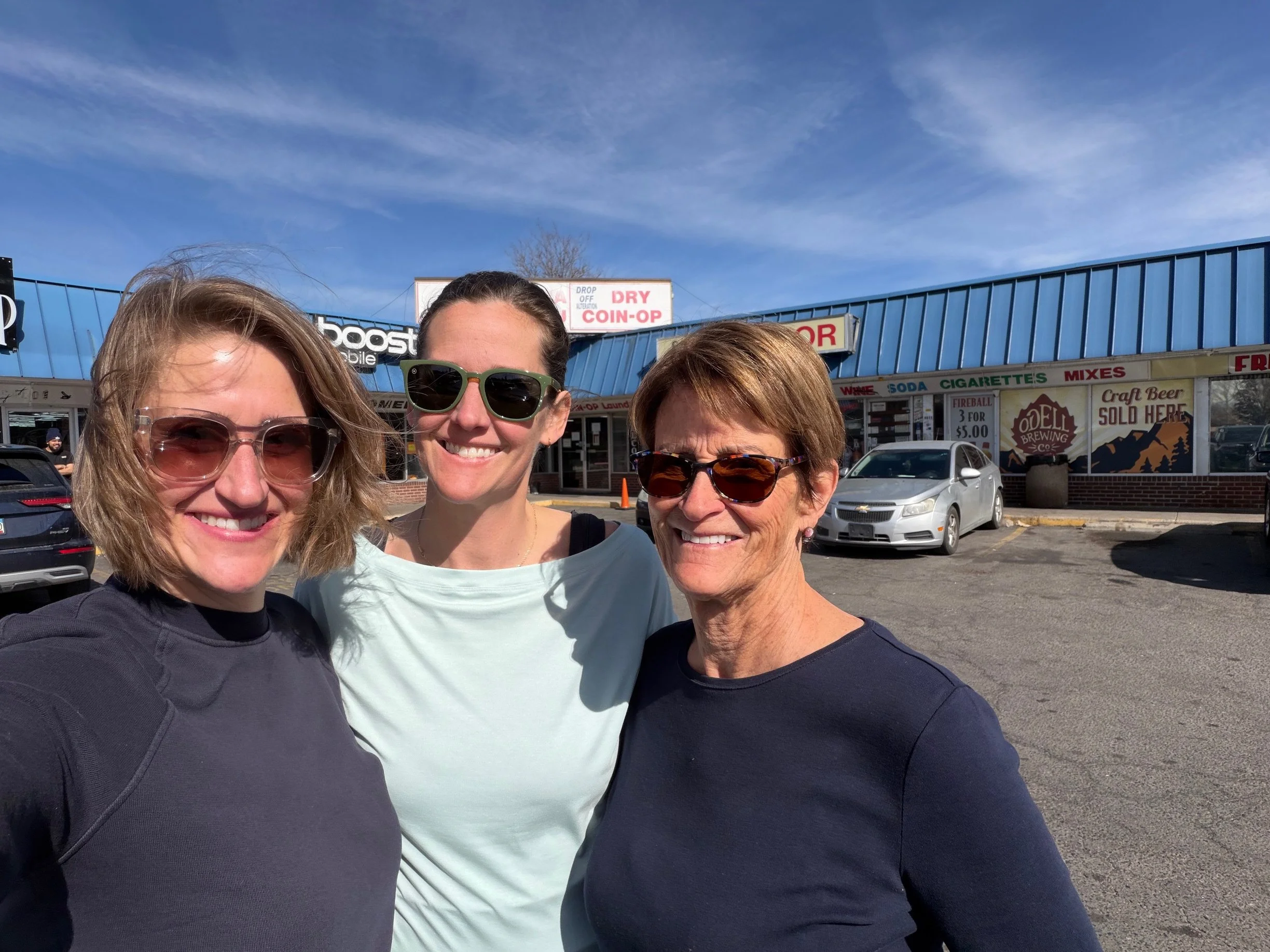 Three women smiling and posing for a selfie outside a shopping plaza with a bright blue sky in the background.