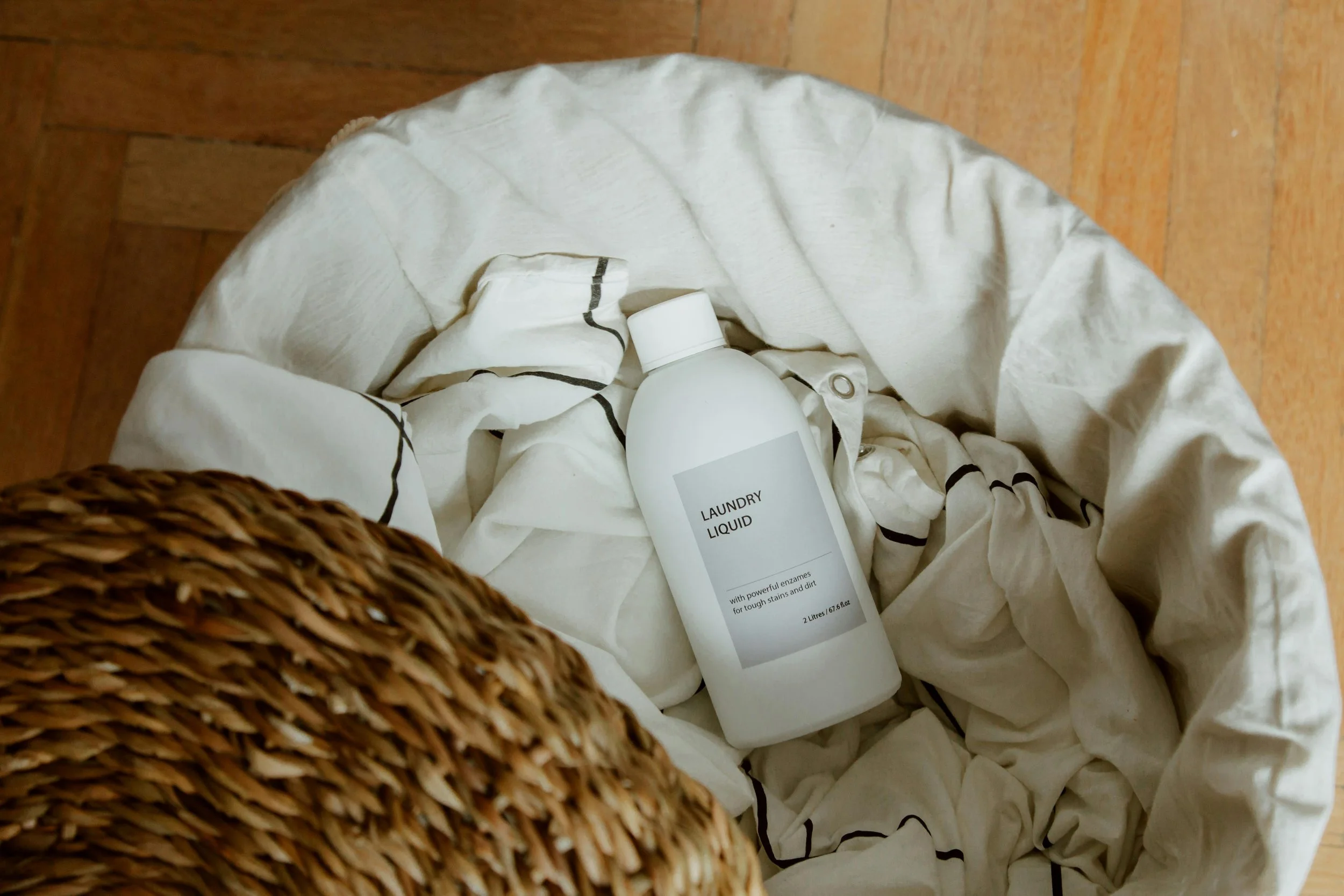 A bottle of laundry liquid placed on a crumpled white fabric with black lines, inside a white laundry basket on a wooden floor.