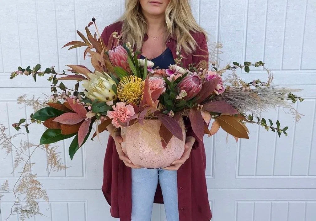 A woman holding a large, colorful floral arrangement in a pink stone bowl, standing in front of a white panel door.