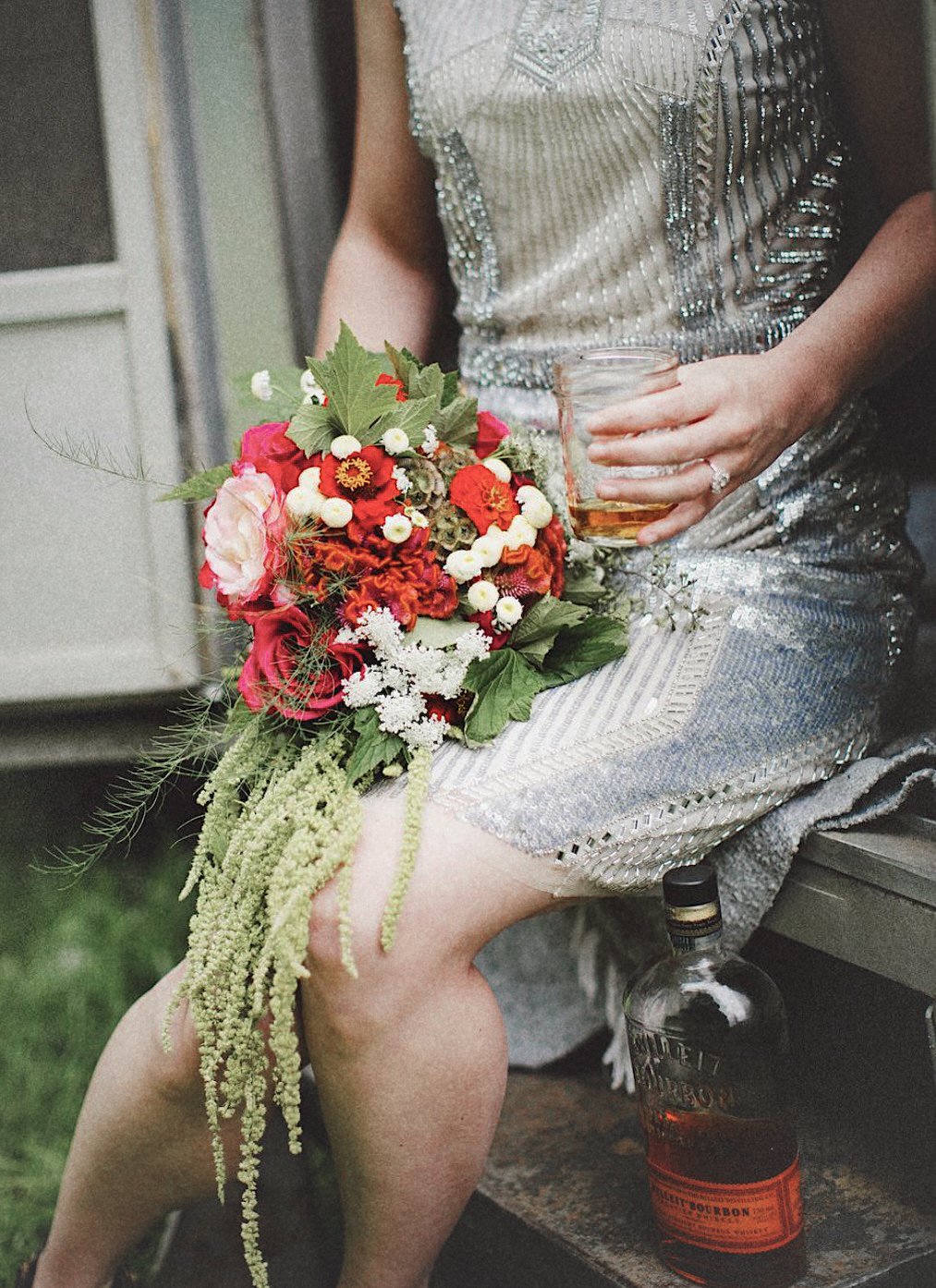A woman in a silver, sparkly dress sitting on a bench, holding a glass of whiskey or similar drink, with a bouquet of red, white, and pink flowers resting on her lap, and a bottle of bourbon on the ground beside her.