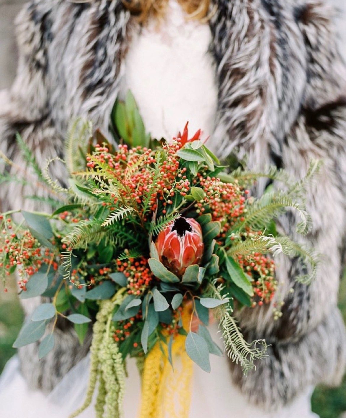 Person holding a bouquet of various flowering plants and green foliage, wearing a dress and a fur coat.