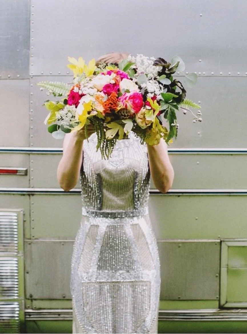 Person in a sparkly silver dress holding a large, colorful bouquet of flowers in front of their face, standing in front of a metallic wall.