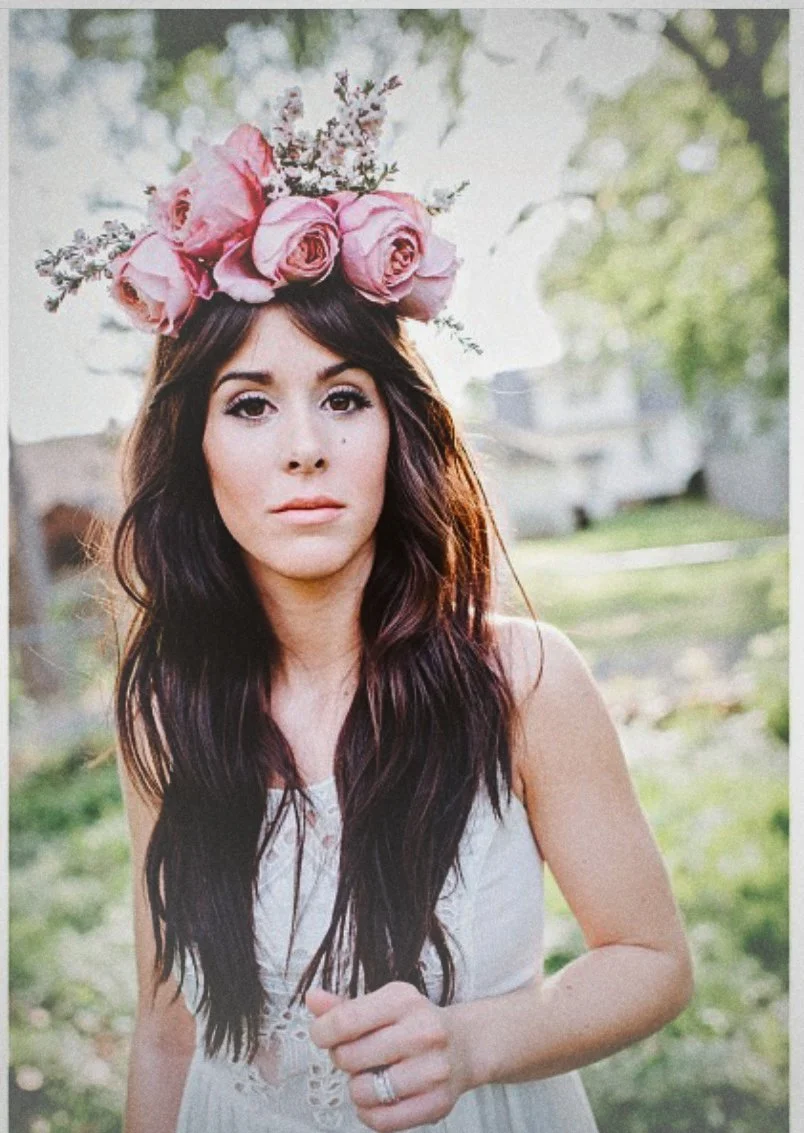 A woman with long dark hair wearing a flower crown of pink roses and smaller flowers, standing outdoors in a green yard.