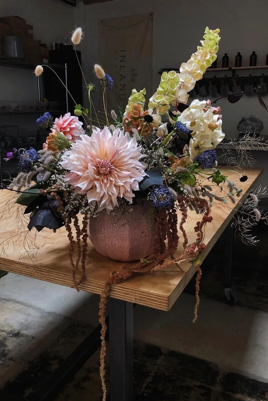 A large floral arrangement in a pink textured vase features various flowers including large peach-colored dahlias, white and yellow snapdragons, purple scabiosa, and tiny white baby's breath, with some dried brown accents, arranged on a wooden table 
