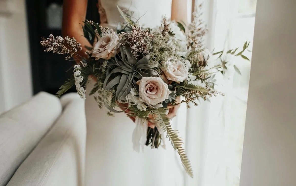 Person holding a bouquet of cream roses, white filler flowers, and green foliage near a window.