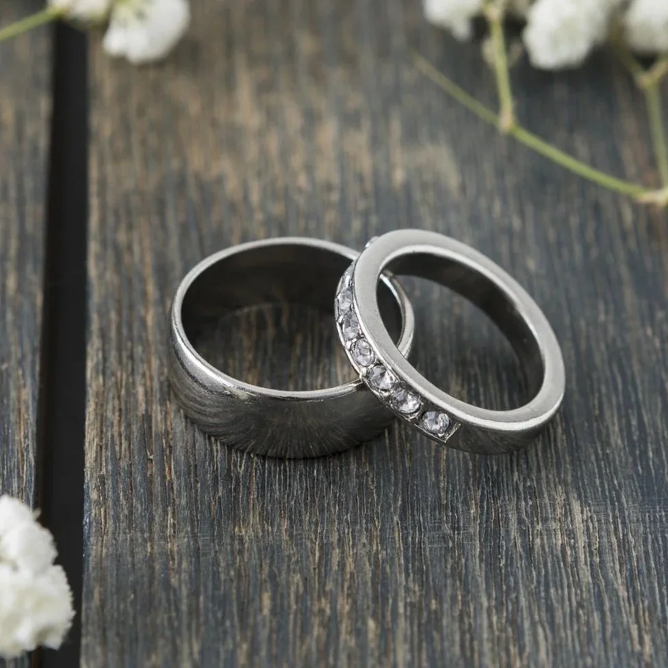 Two silver rings, one plain and one with embedded clear gemstones, are placed on a dark wooden surface with some white flowers in the background.