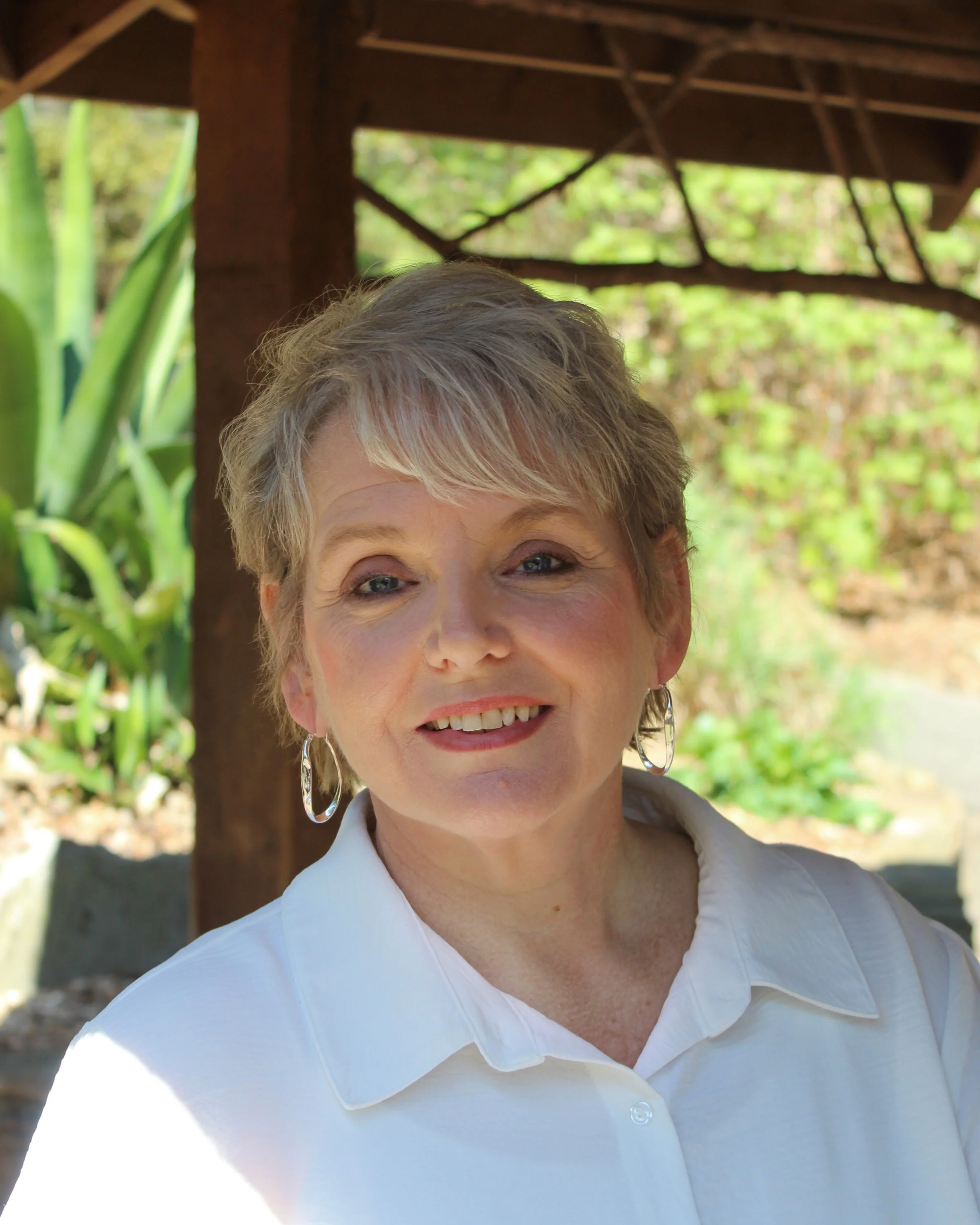 A woman with short blonde hair, wearing a white shirt and hoop earrings, smiling outdoors with green foliage and a wooden structure in the background.