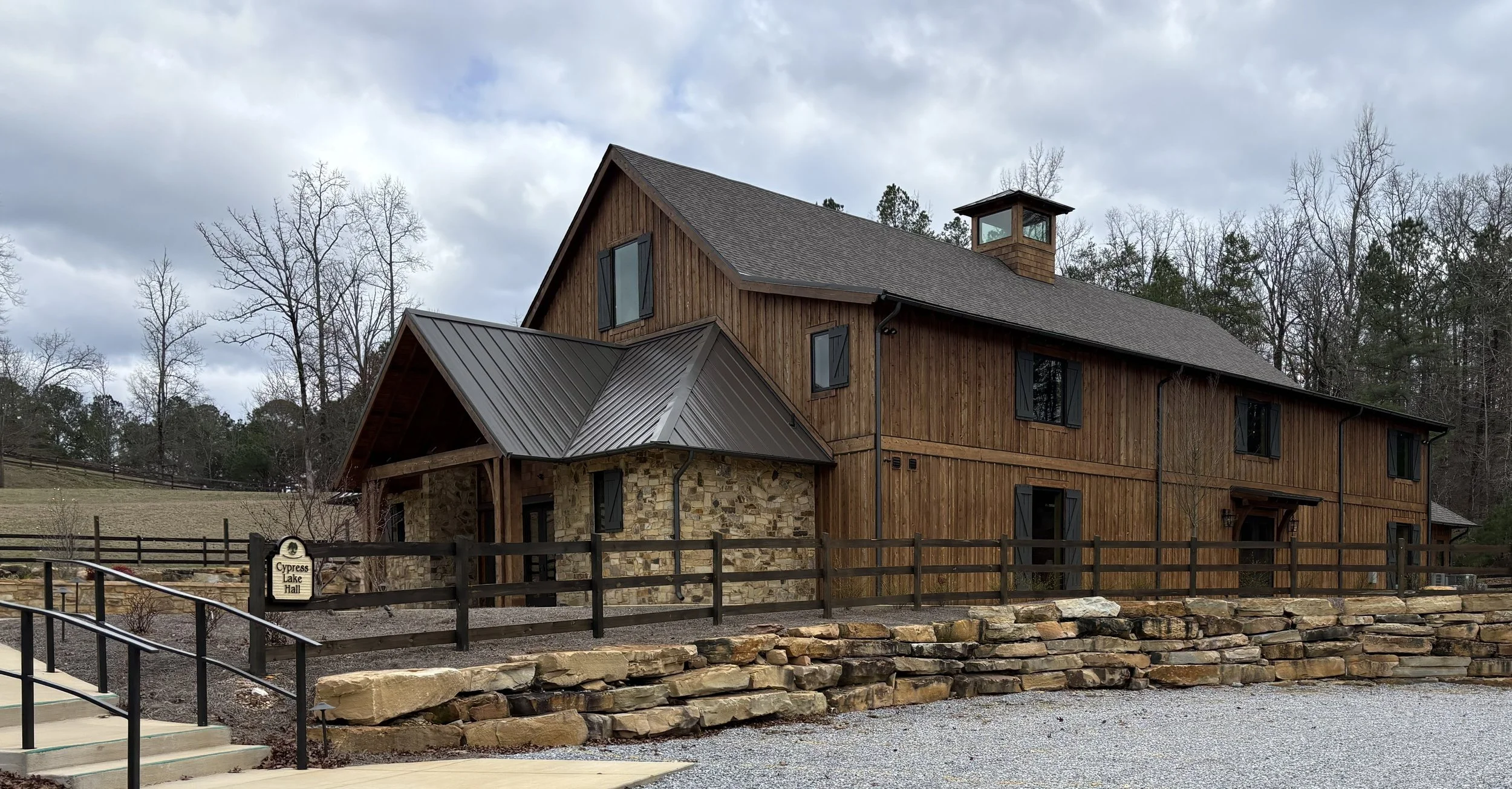 A wooden barn with a stone foundation, multiple windows with black shutters, and a metal roof, surrounded by bare trees and a gravel area.